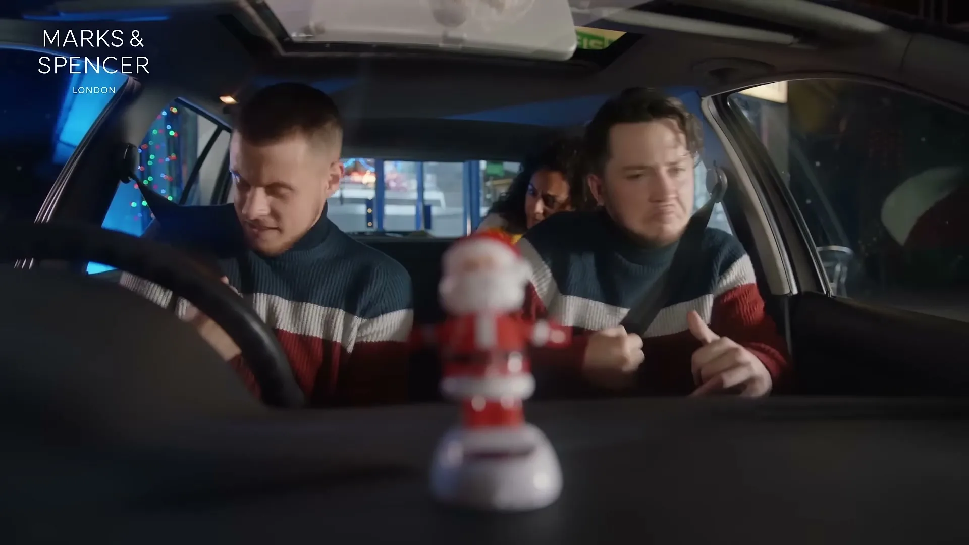 Two men in matching Christmas jumpers and a woman in the back seat are in a car at night, with a bobblehead Santa on the dashboard.