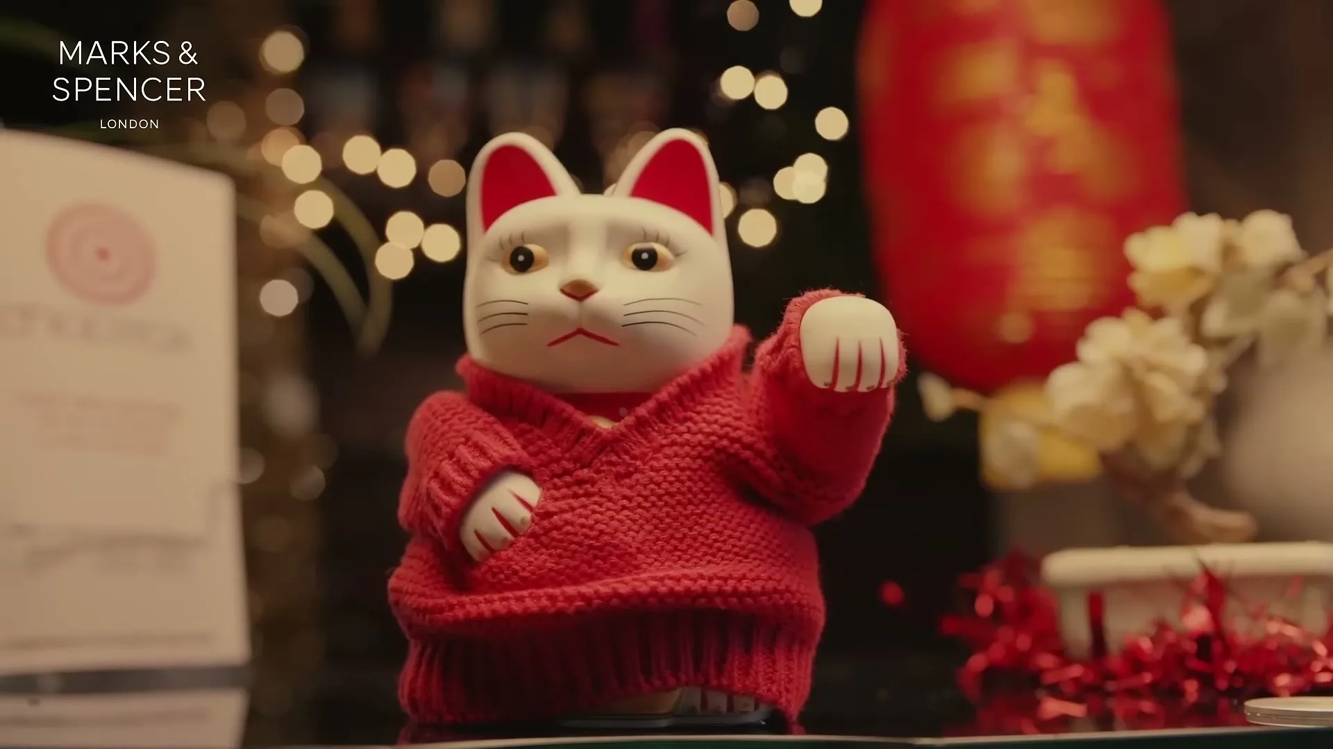 A white Maneki-neko cat figurine wearing a red knitted jumper waves its paw on a reflective surface, with festive lights and a red lantern in the blurred background.