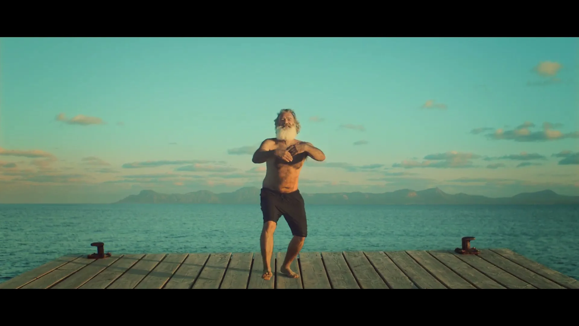 A shirtless, bearded man with a neck brace dances on a wooden pier, with the ocean and mountains in the background under a clear sky.