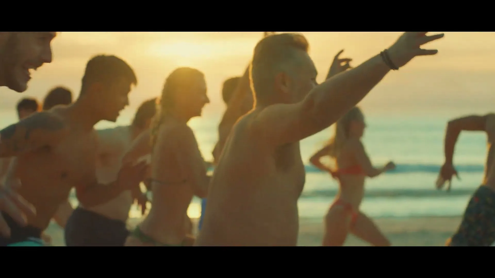 A diverse group of people, mostly in their 20s-40s, run joyfully towards the ocean on a beach at golden hour.