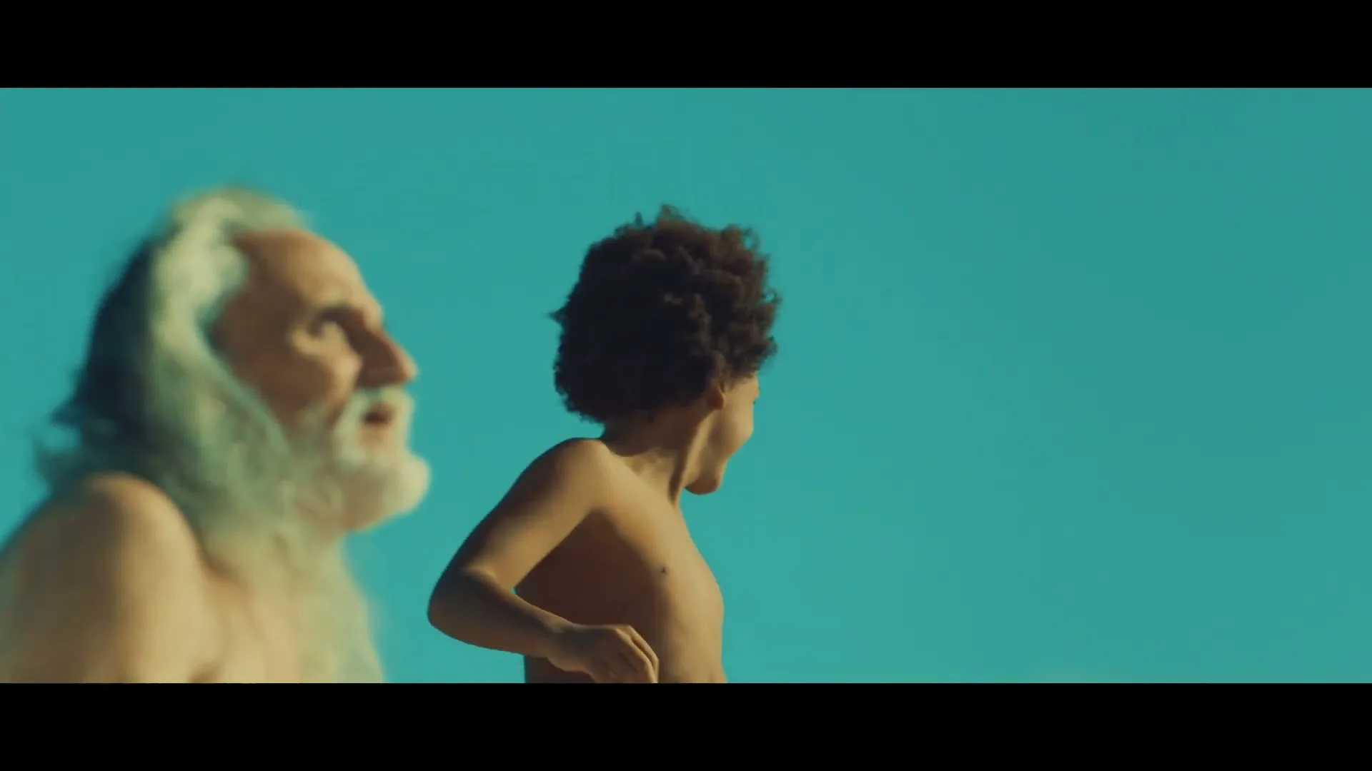 A diverse group of people, including an older man with a long white beard and a young person with curly hair, run joyfully on a sunny beach.