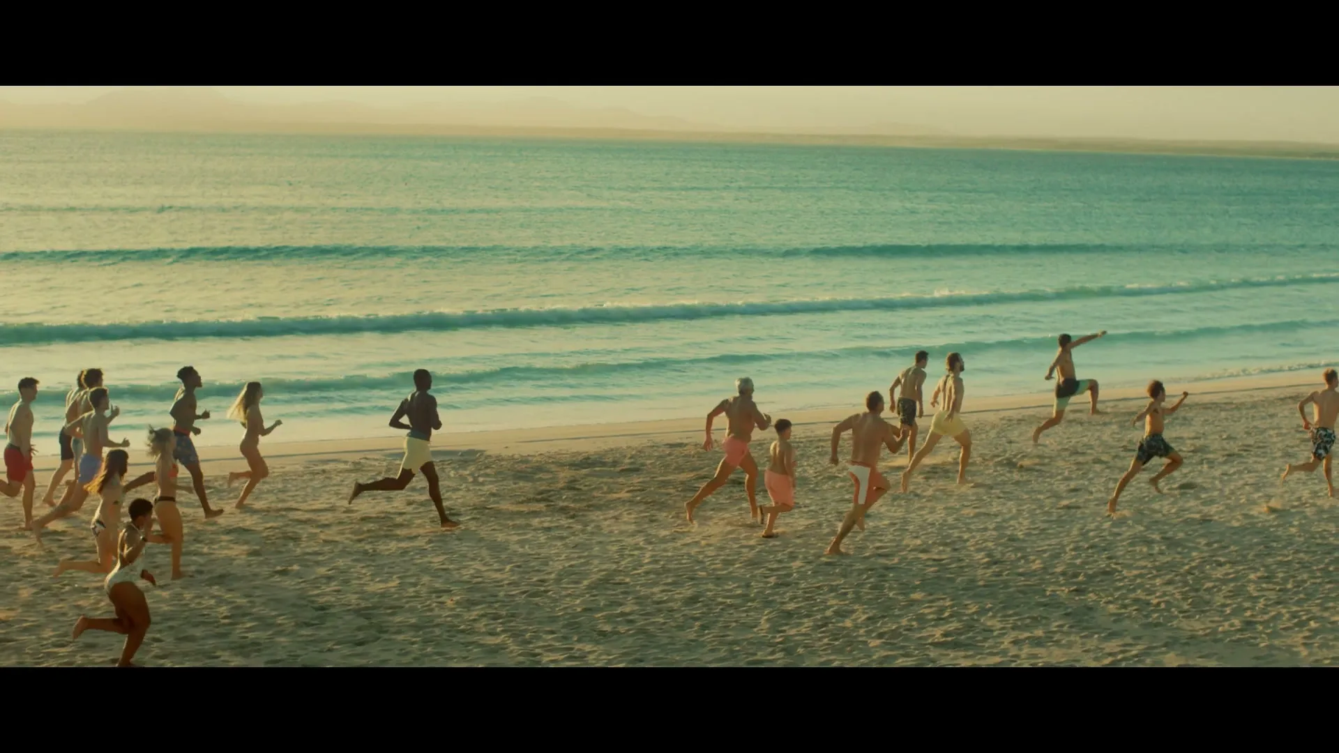 A diverse group of young adults in swimwear runs joyfully along a sandy beach towards the turquoise ocean under a golden sky.