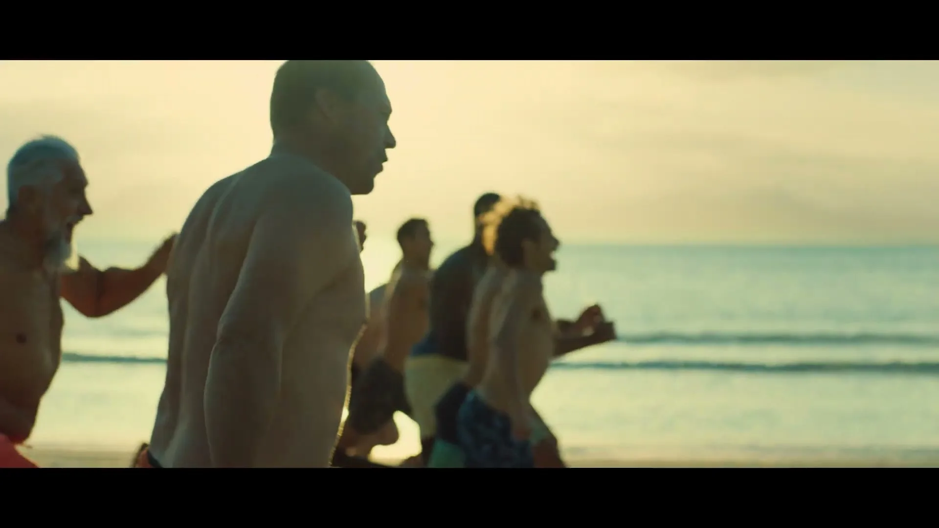 A group of diverse men, shirtless and wearing swim trunks, run joyfully along a sunny beach with the ocean in the background during golden hour.