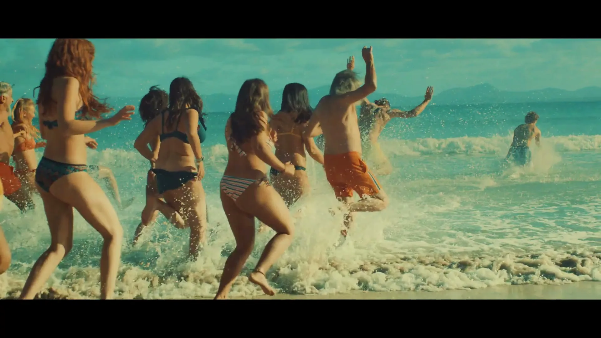 A diverse group of young adults in swimsuits joyfully run into the ocean on a sunny day at a tropical beach.