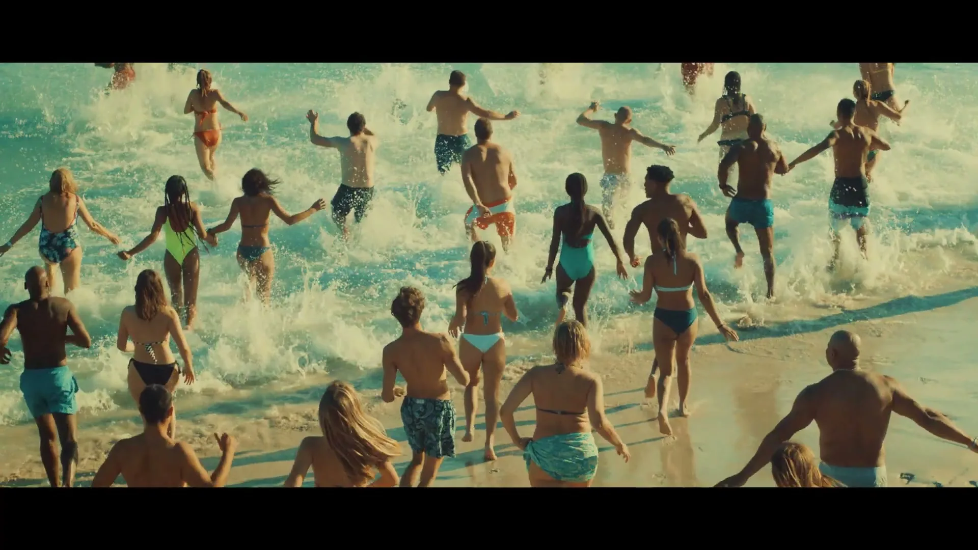 A diverse group of people in swimsuits run into the ocean, splashing through the waves on a sunny day at the beach.