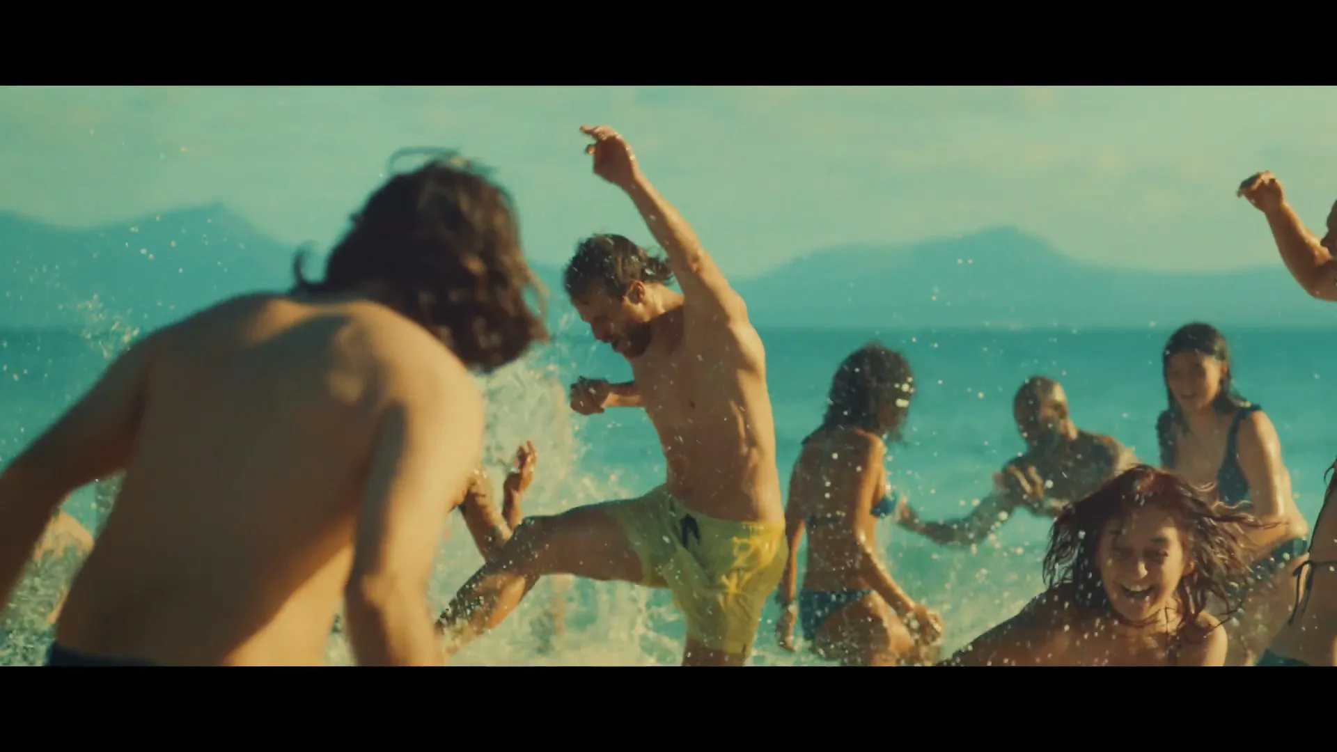 A diverse group of young adults in swimsuits splash and play in the shallow turquoise ocean water on a sunny day at a beach.