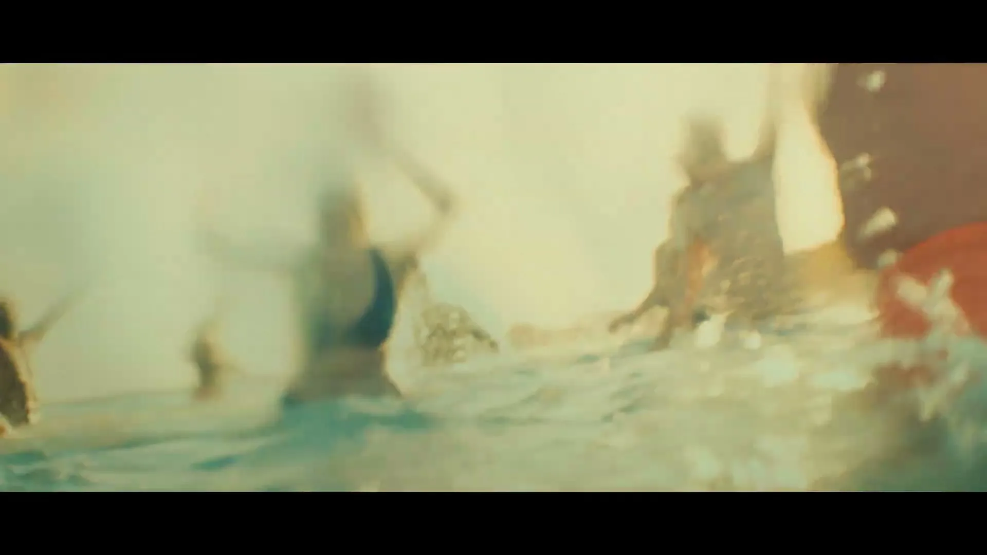 A group of diverse people, some shirtless, some in swimwear, are seen in slow motion, jumping and splashing in the ocean water, with bright sunlight in the background.
