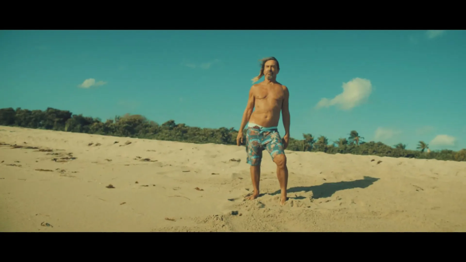 Iggy Pop, shirtless in patterned swim shorts, stands on a sandy beach, looking around under a bright blue sky with scattered clouds.