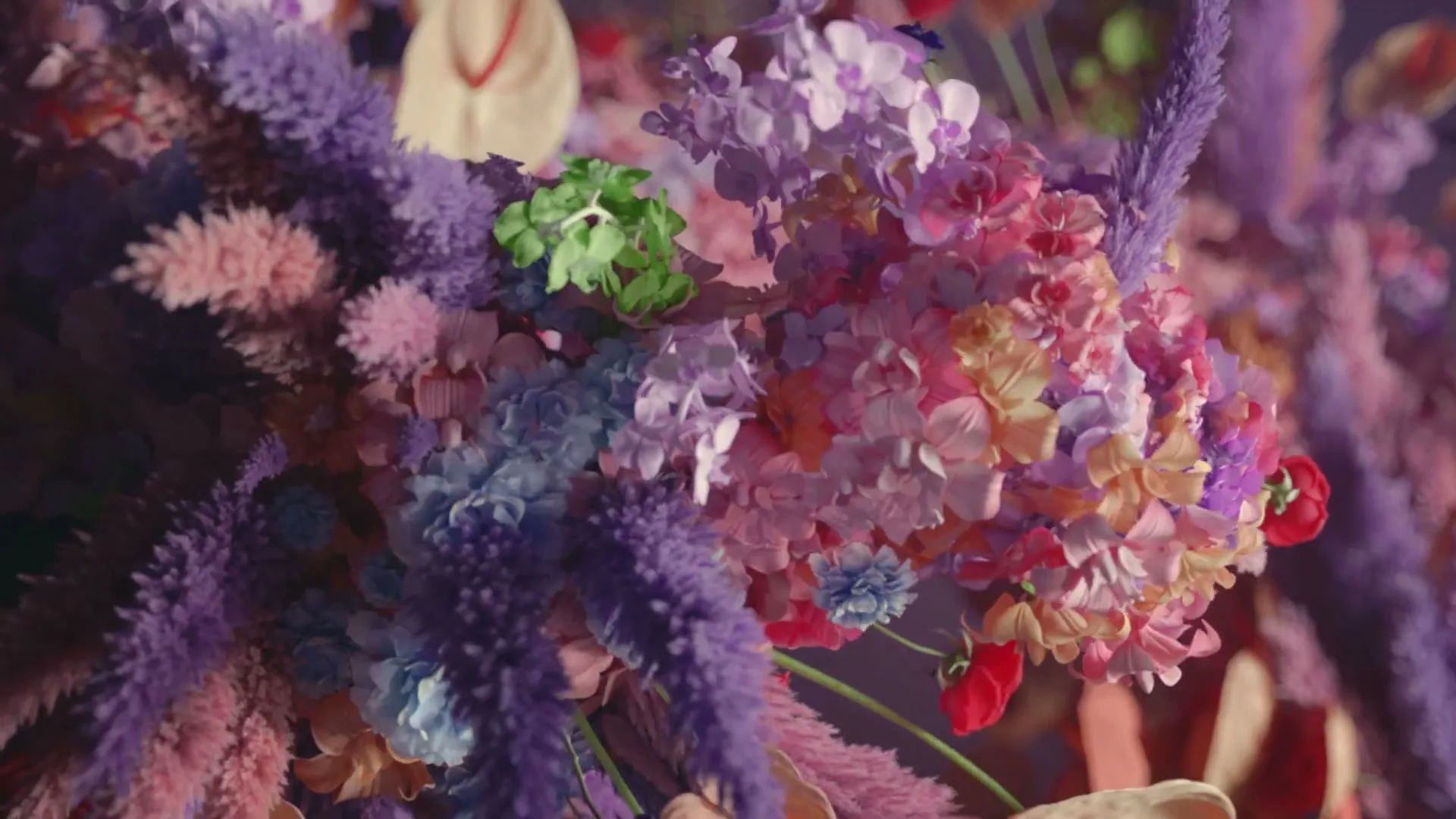 A vibrant, colorful arrangement of various flowers and plants in shades of purple, pink, and green fills the frame.