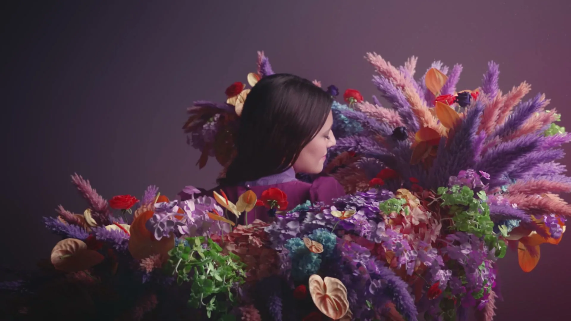 A woman with dark hair, wearing a purple top, is surrounded by a vibrant, colorful arrangement of flowers and plants, looking to the right.