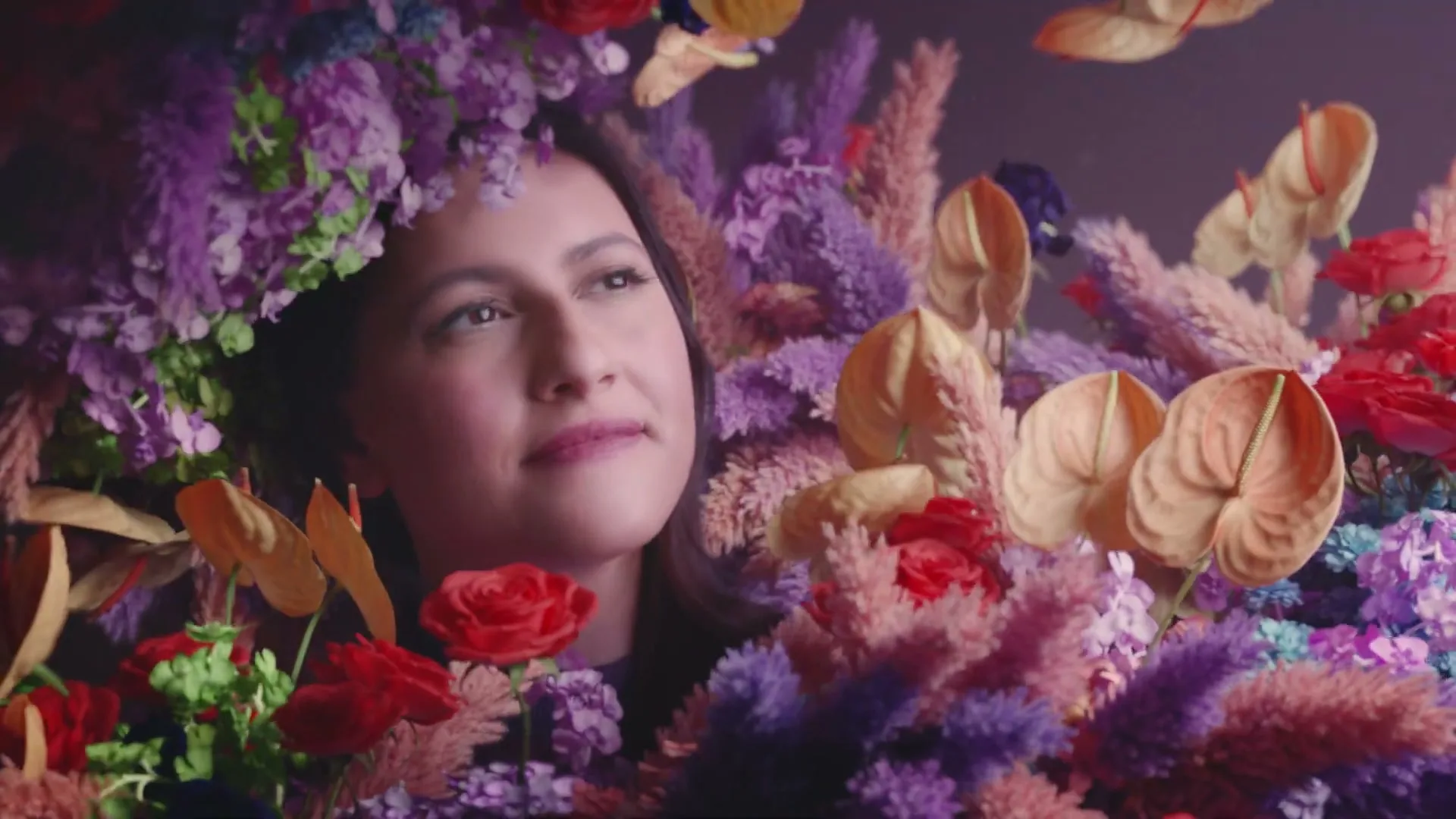A woman with dark hair and closed eyes is surrounded by a vibrant array of purple, red, and orange flowers, with her head tilted slightly upwards.