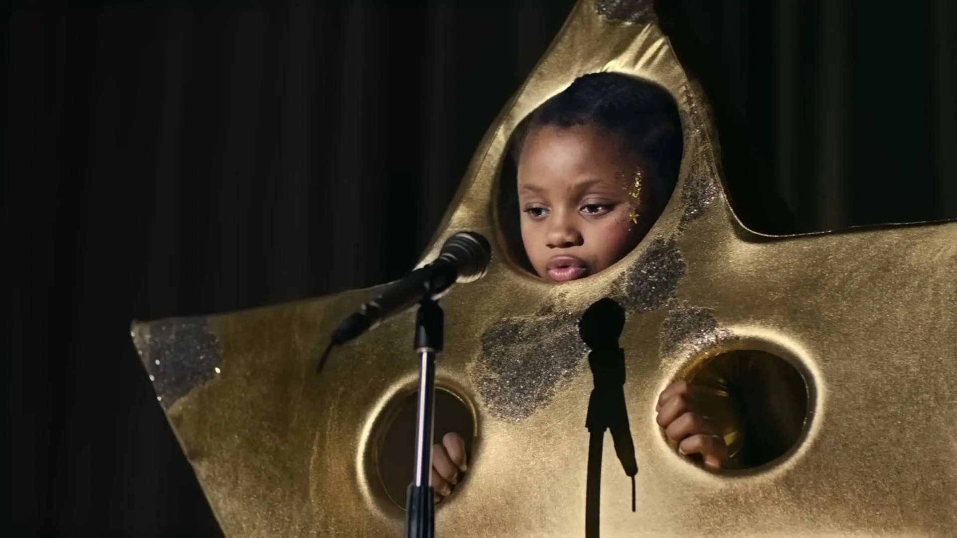 A young girl in a gold star costume with glitter on her face sings into a microphone on a dark stage with a black curtain.