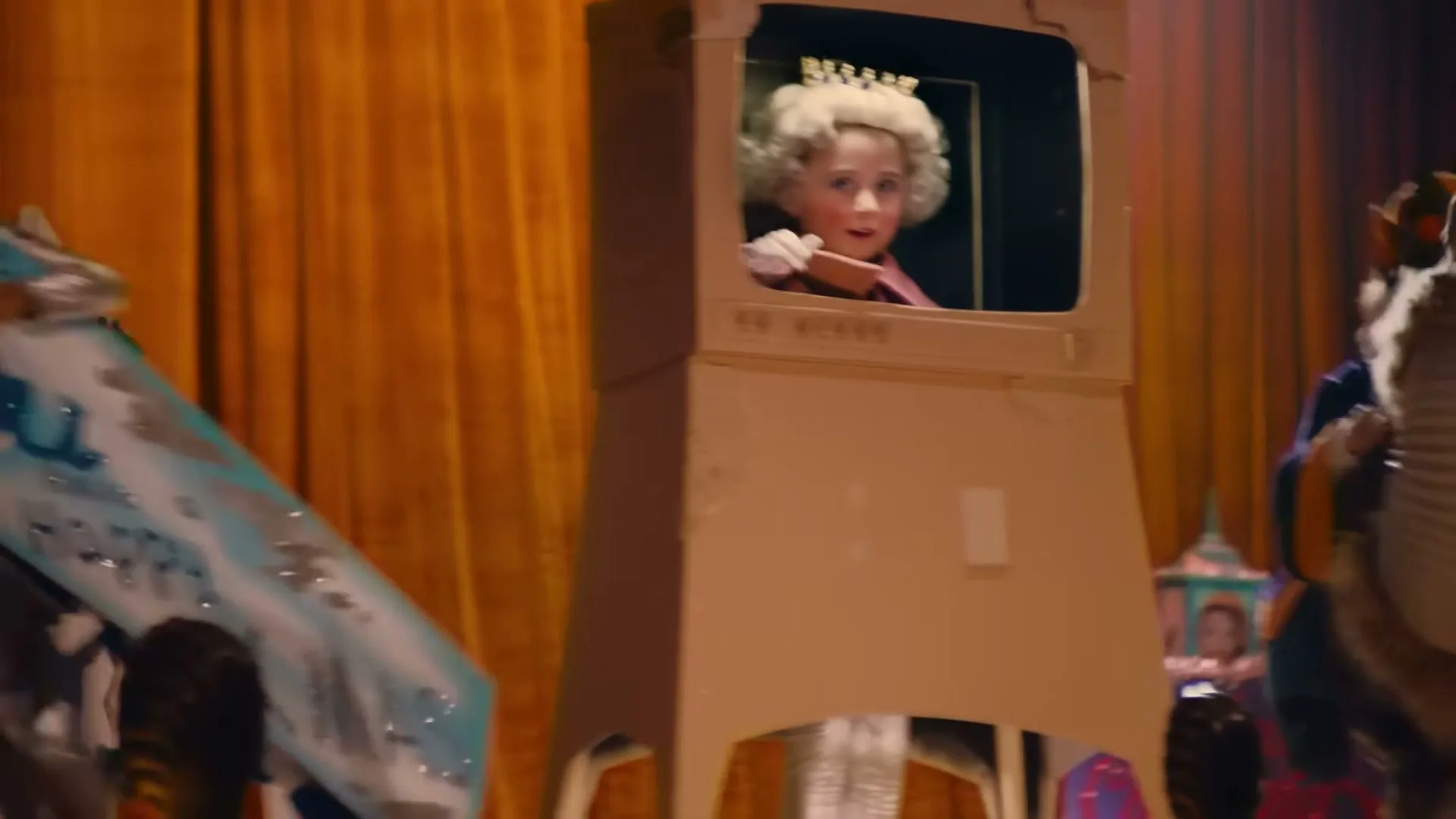 A young girl dressed as Queen Elizabeth II, wearing a blonde wig and crown, waves from inside a cardboard television set on a stage with orange curtains.