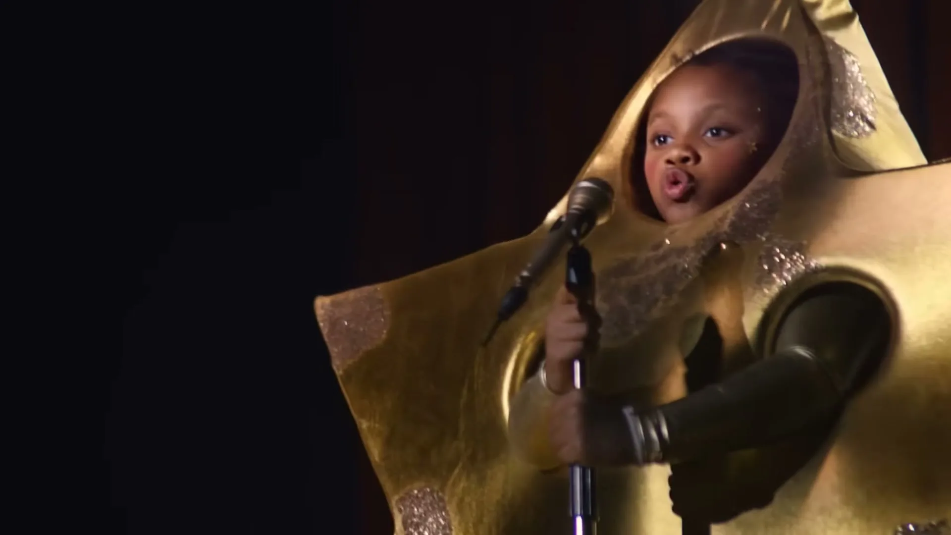 A young Black girl in a gold cheese costume sings into a microphone on a dark stage, looking determined.