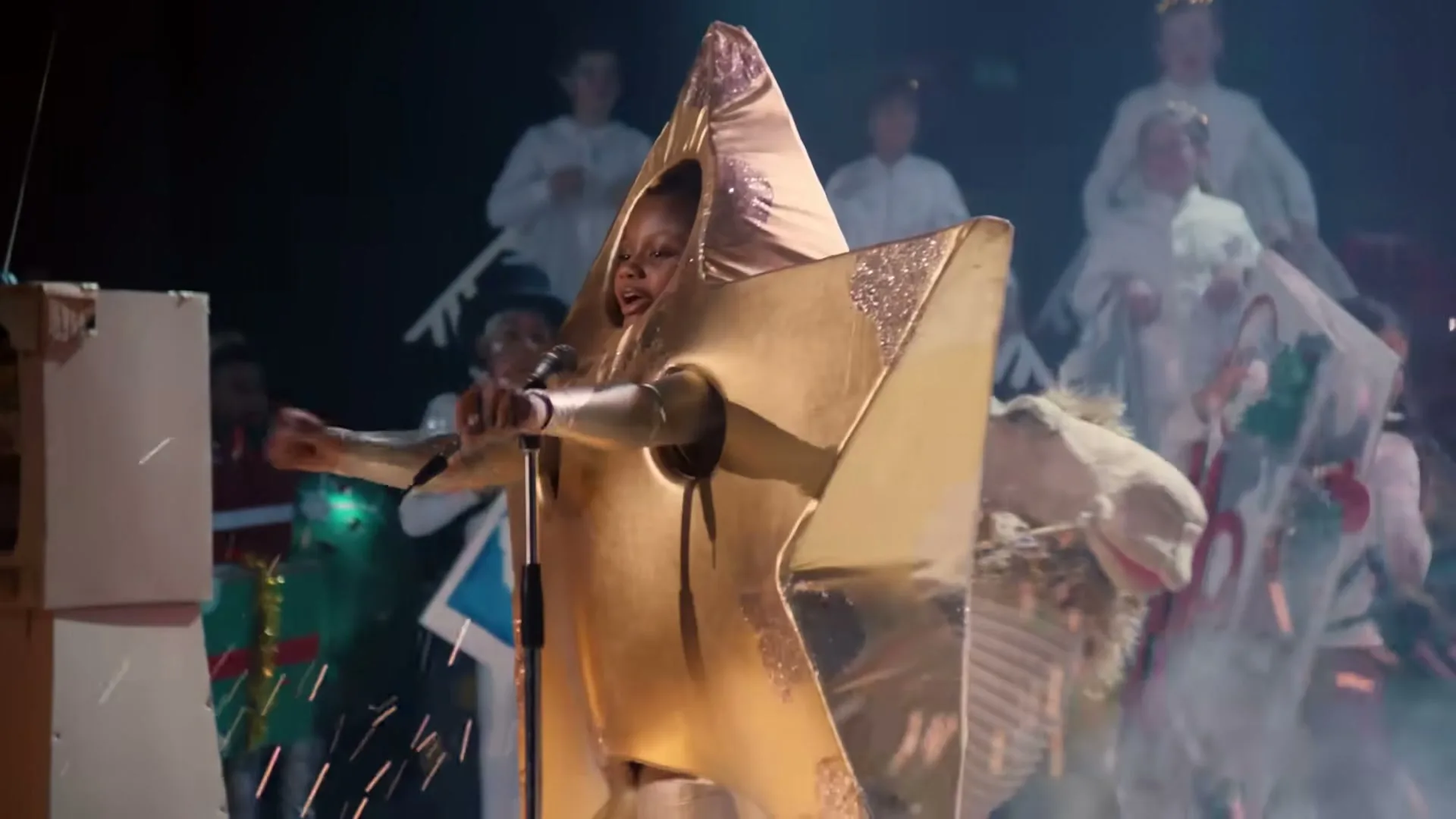 A young girl in a gold star costume sings into a microphone on stage, surrounded by other children in various costumes, with sparks flying around her.