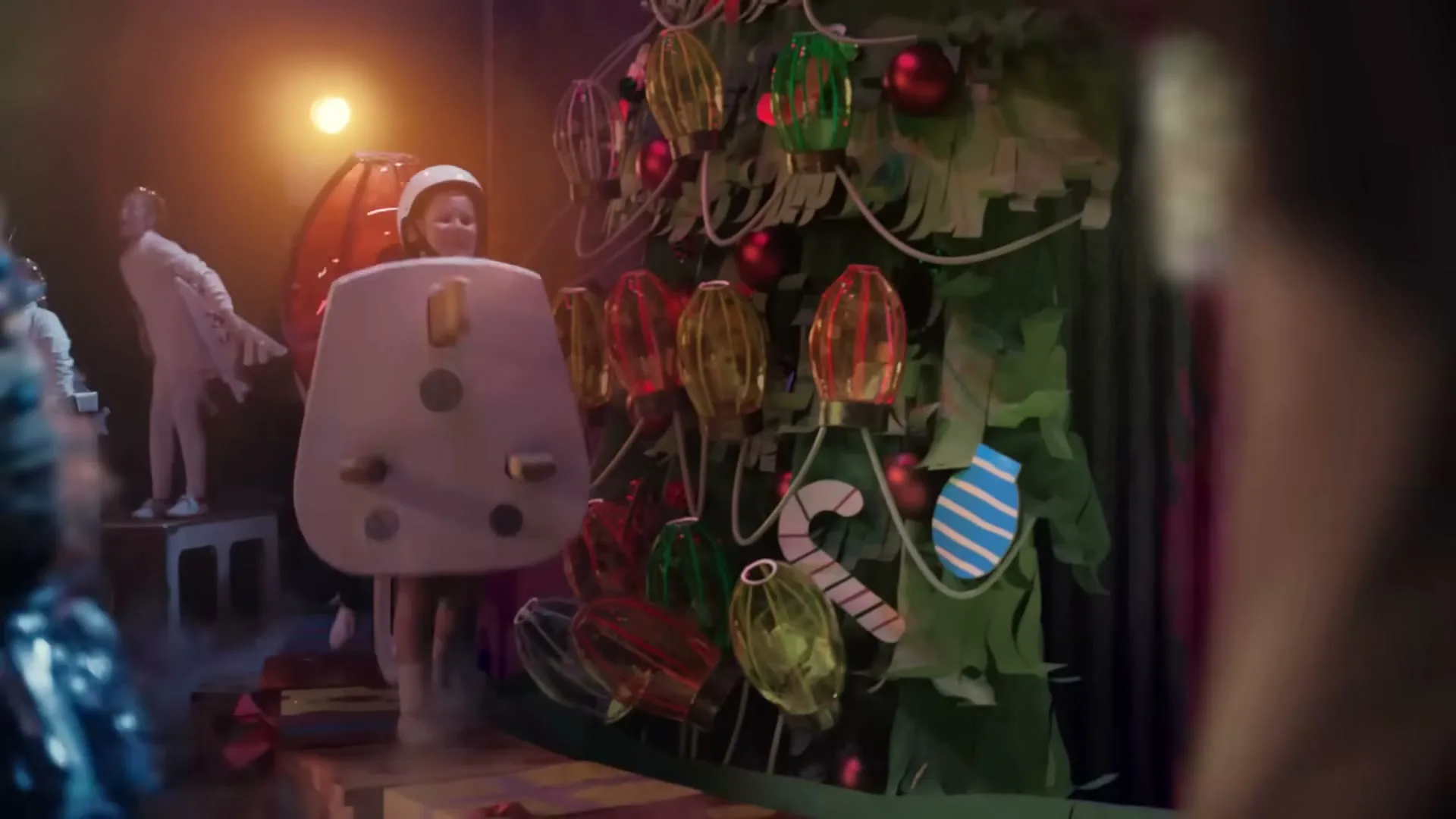 A child dressed as a large electrical plug walks on a stage with other children in costumes, in front of a Christmas tree prop.
