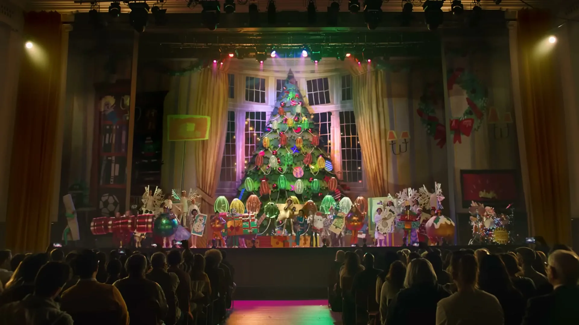 A wide shot of a school play with children dressed as Christmas ornaments on a stage, performing in front of an audience.