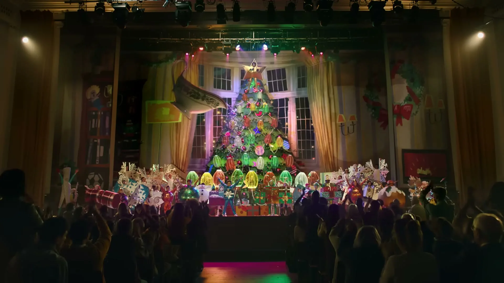A wide shot of a school play with children dressed as food items on stage, performing in front of an audience.