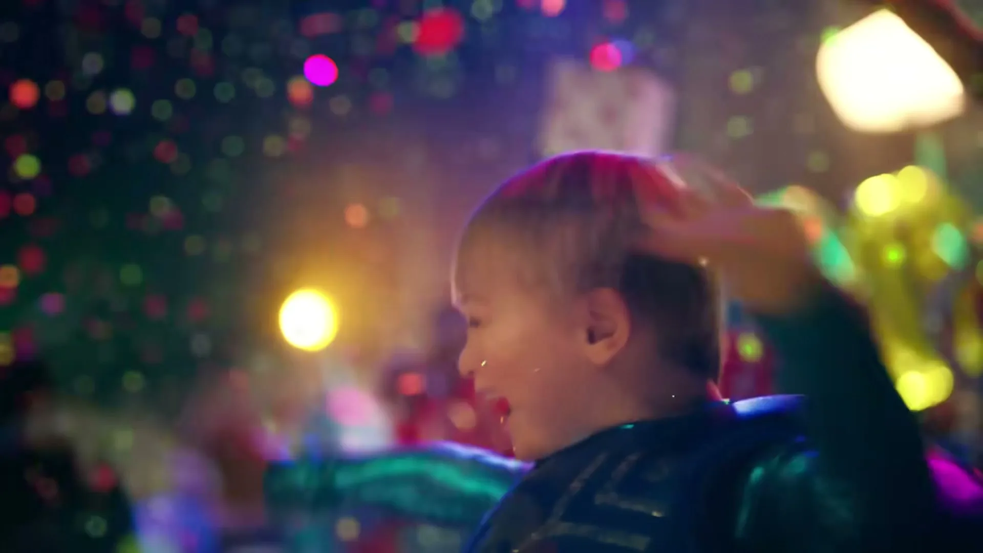 A young child with blonde hair, wearing a blue costume, dances joyfully as colorful confetti falls around them in a vibrant, festive interior setting.