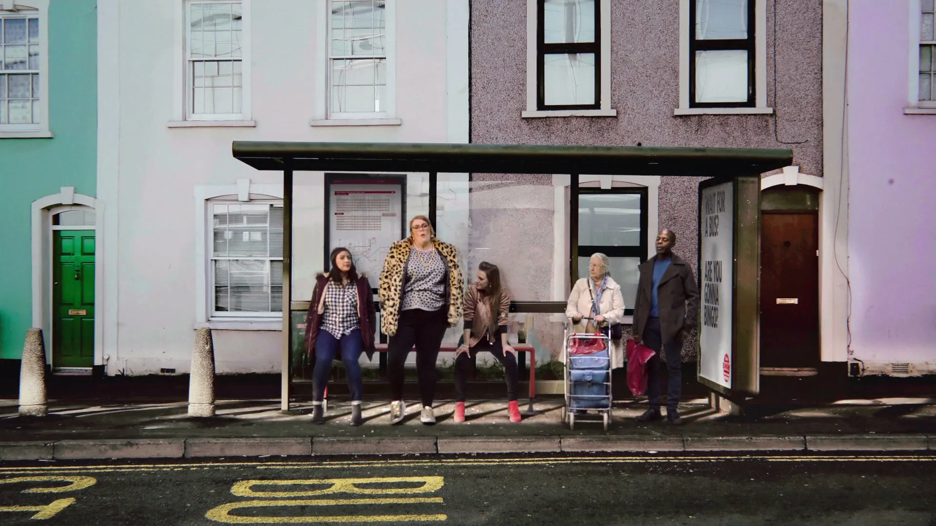Five diverse people wait at a bus stop in front of colorful terraced houses, as a car drives past, obscuring the view.