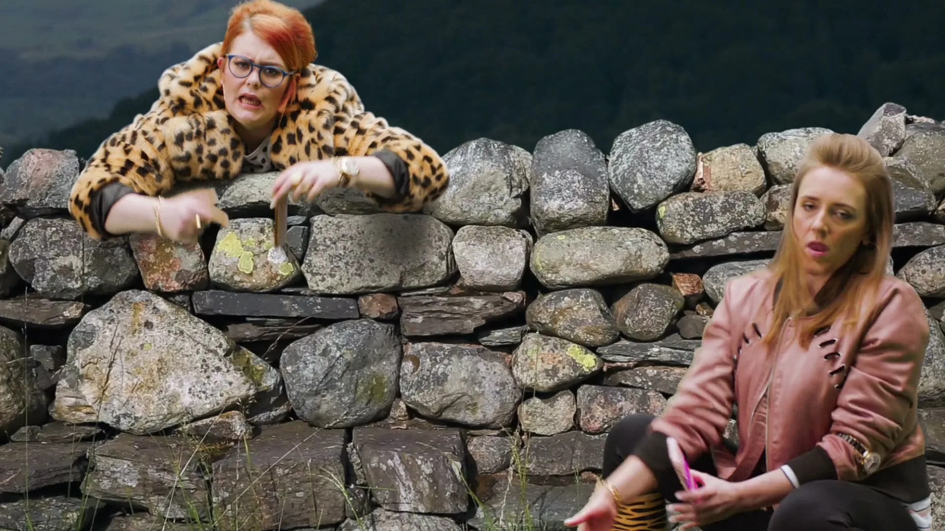 Two women in animal print clothing pose with their phones in front of a stone wall, pointing at the screens.