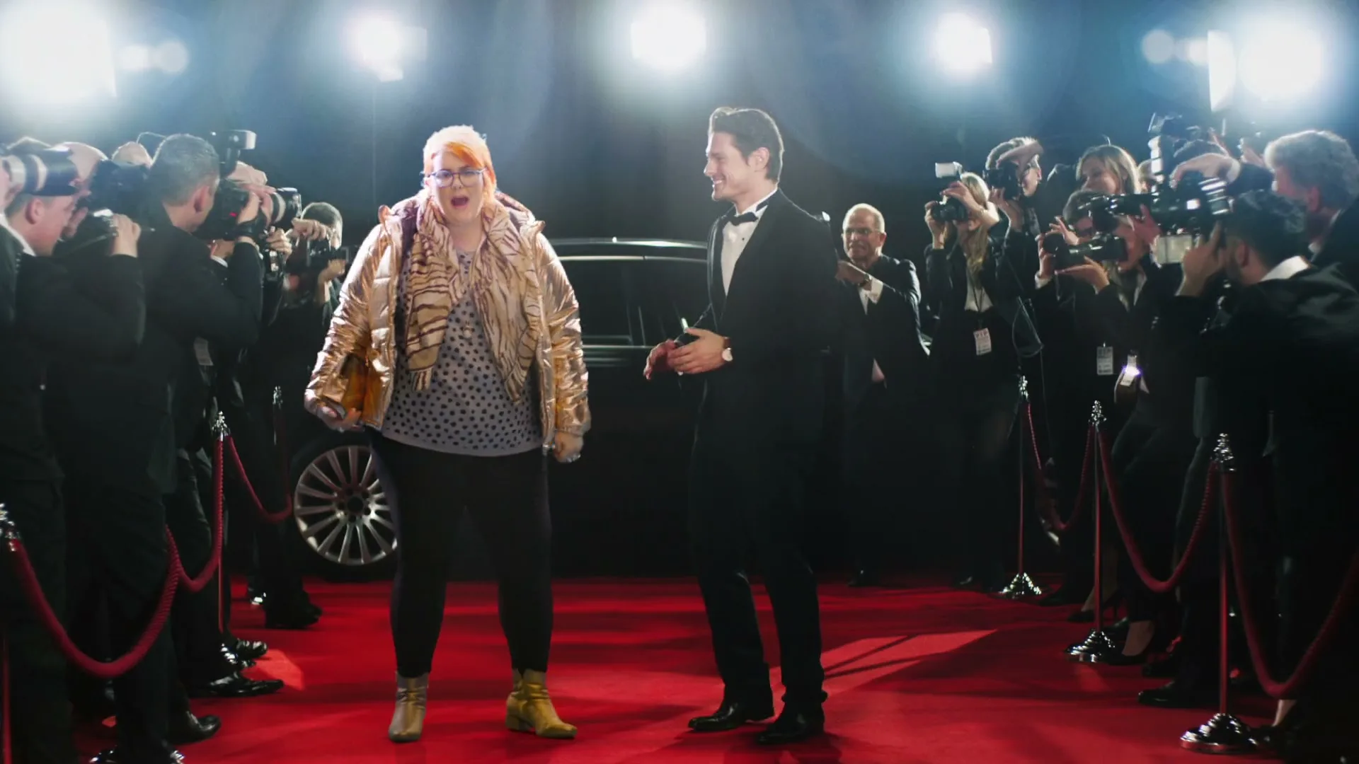 A woman in a gold jacket and a man in a tuxedo walk down a red carpet, surrounded by paparazzi and bright camera flashes.