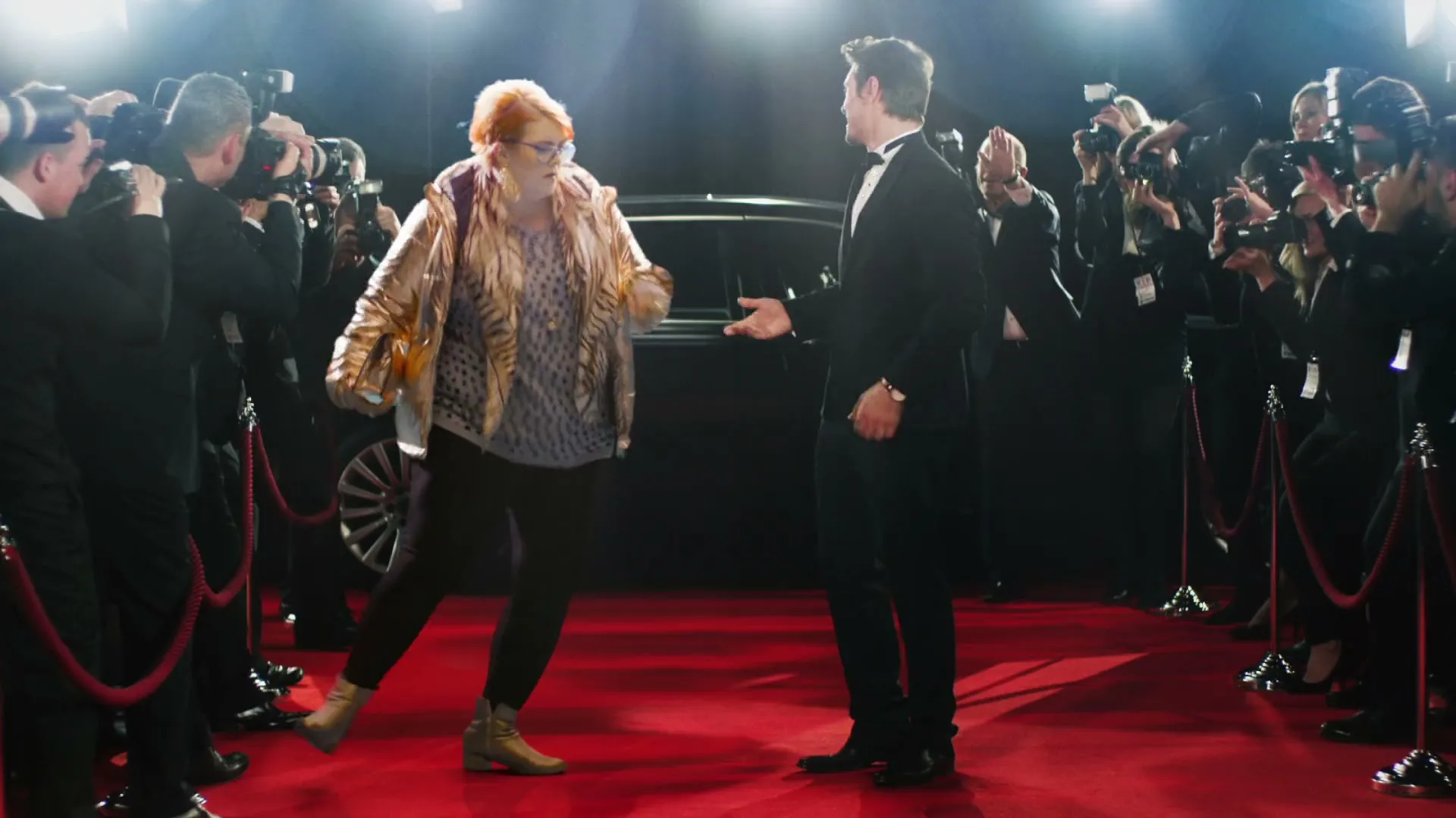 A woman in a gold jacket dances on a red carpet, surrounded by paparazzi and a man in a tuxedo, with a black car in the background.