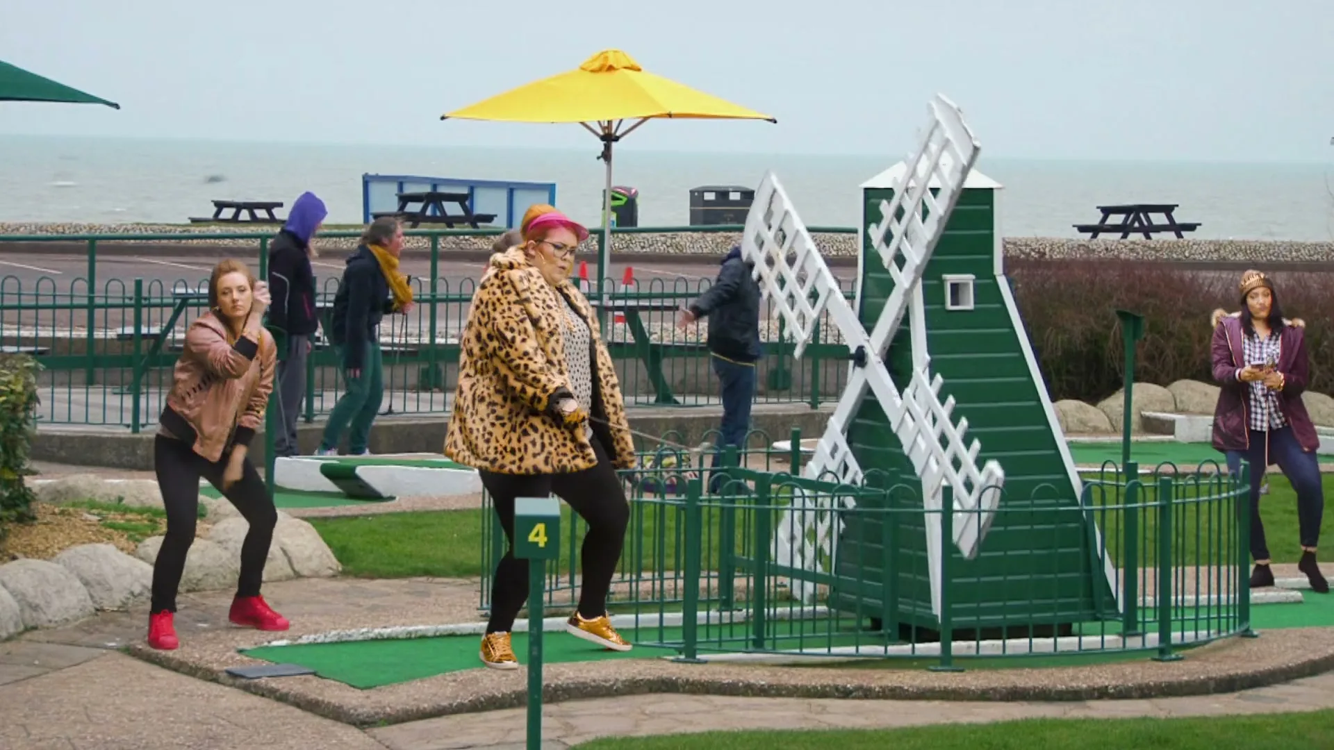 A diverse group of people dance and play mini golf at a seaside course, with a woman in a leopard print coat at the center.