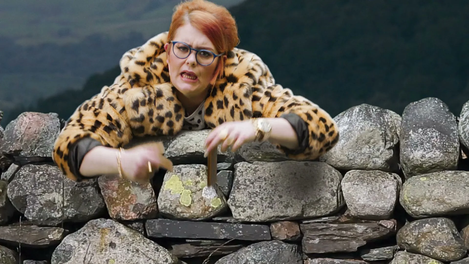 A woman with red hair and glasses, wearing a leopard print coat, leans over a stone wall, holding up a phone with the Sun Bingo logo.