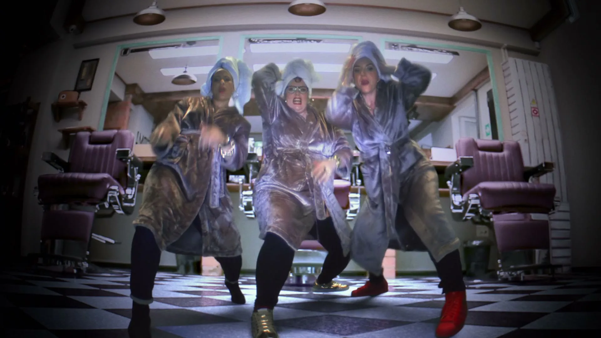 Three women in bathrobes and hair towels dance energetically in a barbershop with checkered floors and vintage chairs.