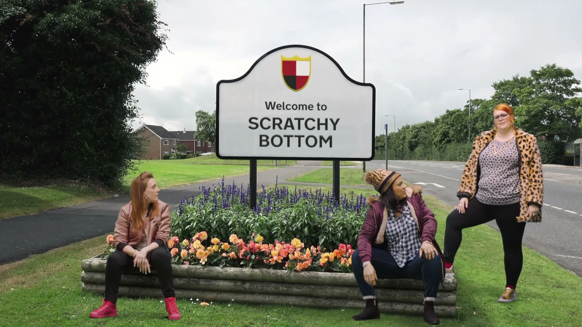 Three women pose and dance in front of a "Welcome to Scratchy Bottom" sign on the side of a road, with cars passing by.