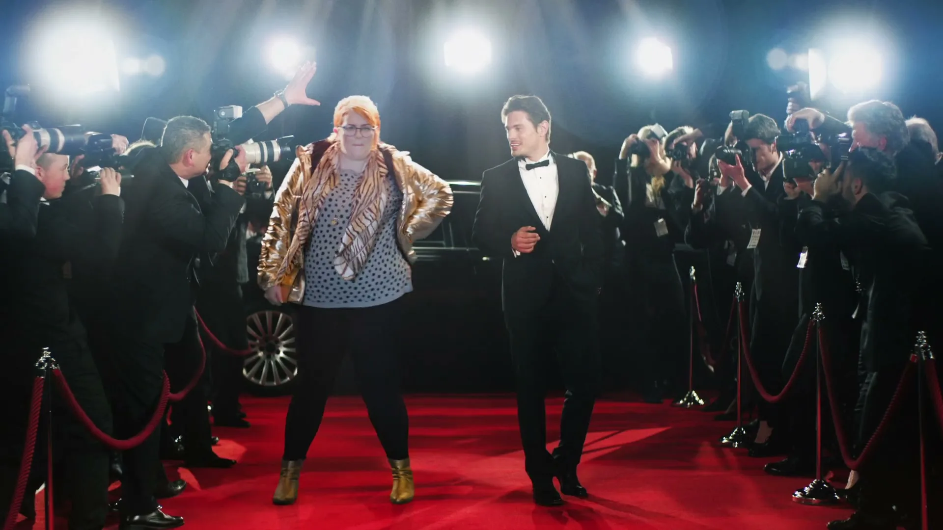 A woman in a gold jacket and a man in a tuxedo walk down a red carpet, surrounded by photographers and bright lights.