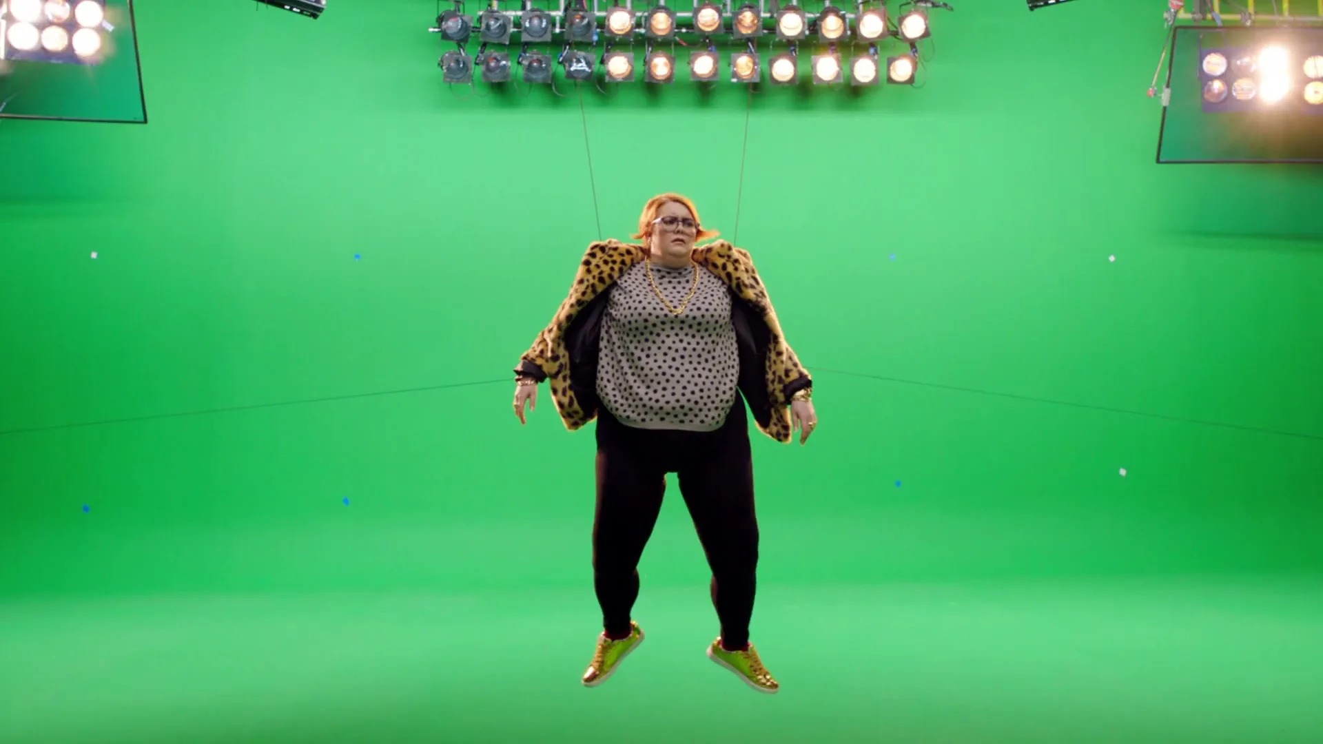 A woman in a leopard print jacket and black leggings is suspended in the air against a green screen, with studio lights visible above.