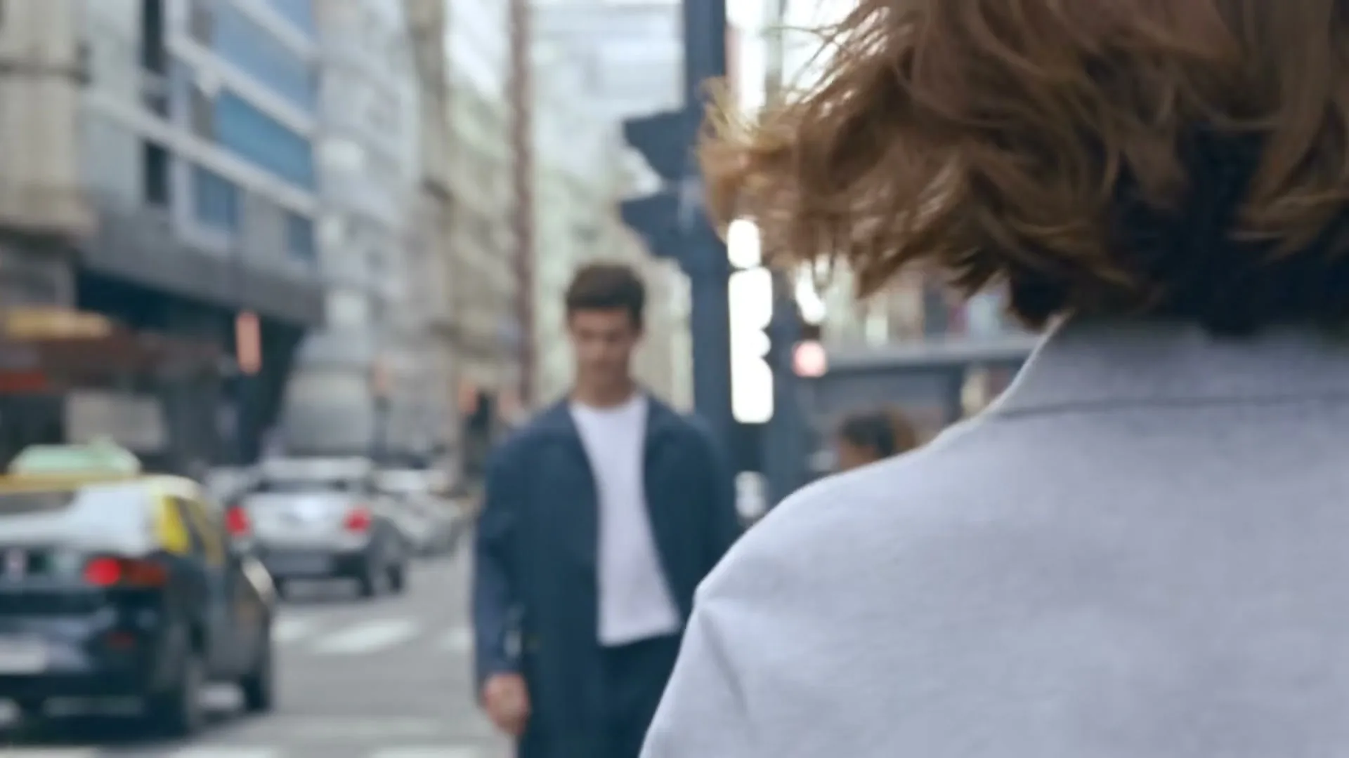 A young man with dark hair and a white t-shirt walks towards the camera on a city street, looking directly at the viewer.