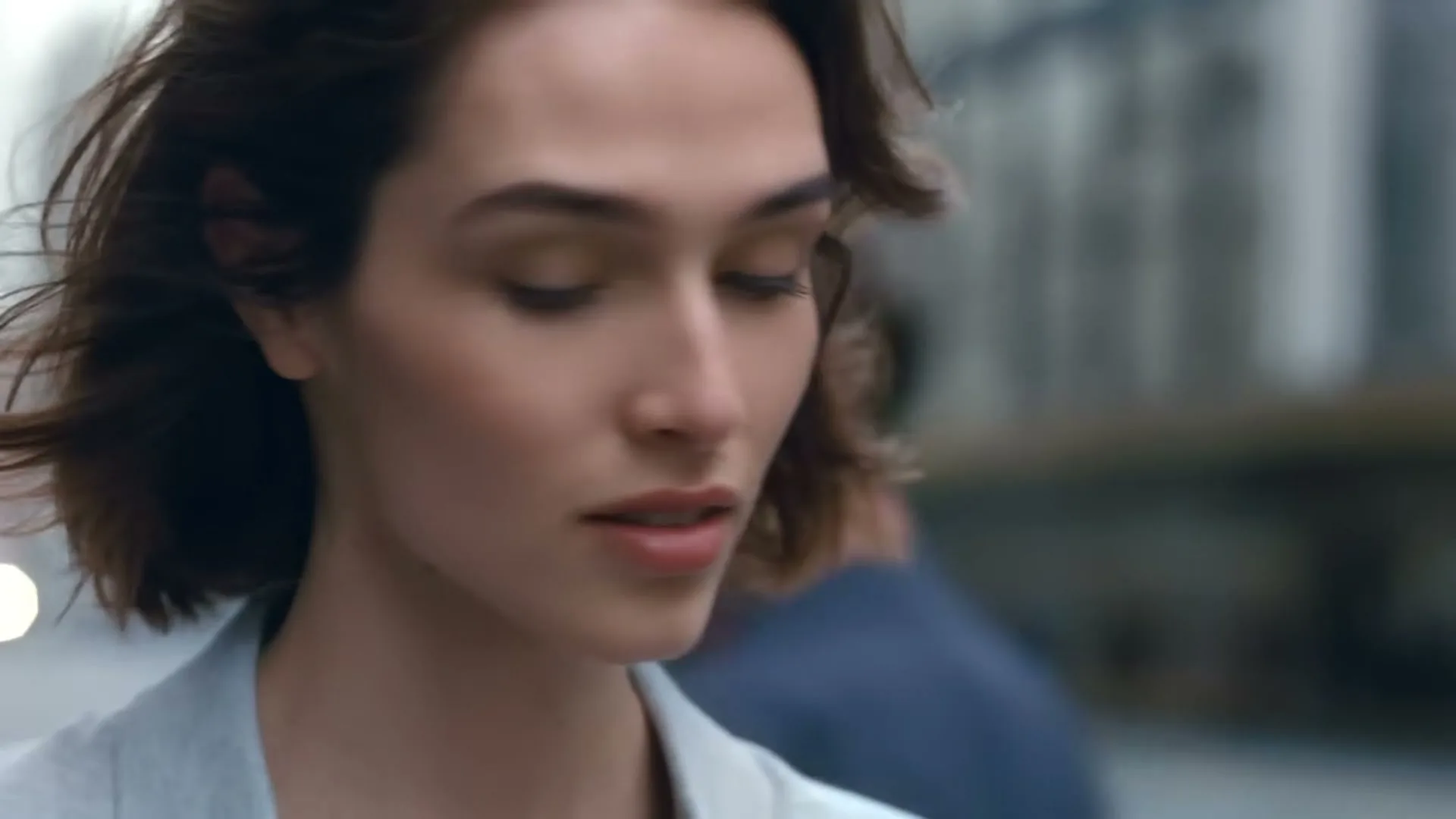 A young woman with short brown hair and a light blue jacket walks forward, looking around a busy city street.