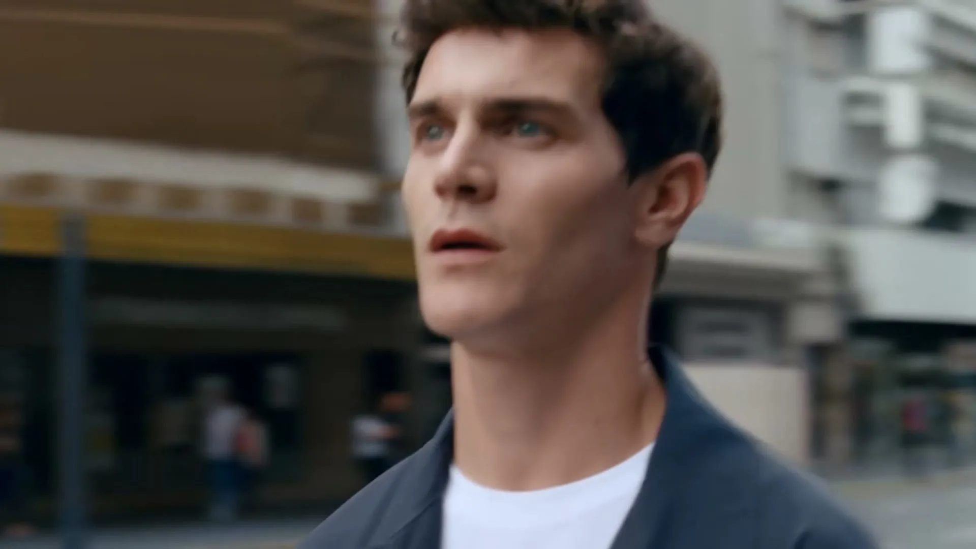A young man with dark curly hair and a dark blue jacket walks down a busy city street, looking around, then turns to look at a woman.