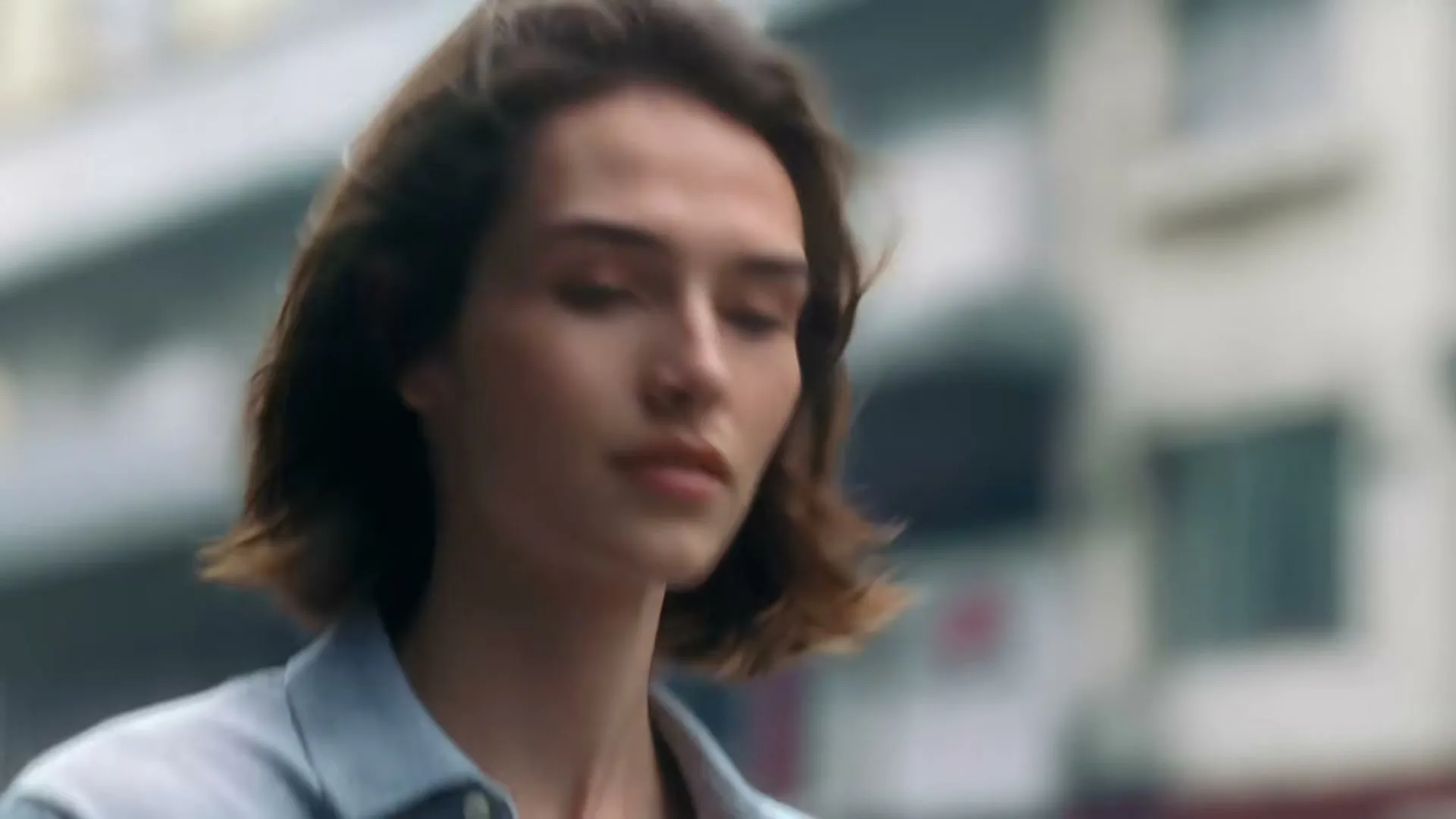 A young woman with short brown hair and a light blue shirt looks to her right, then turns her head to the left, with buildings in the blurred background.