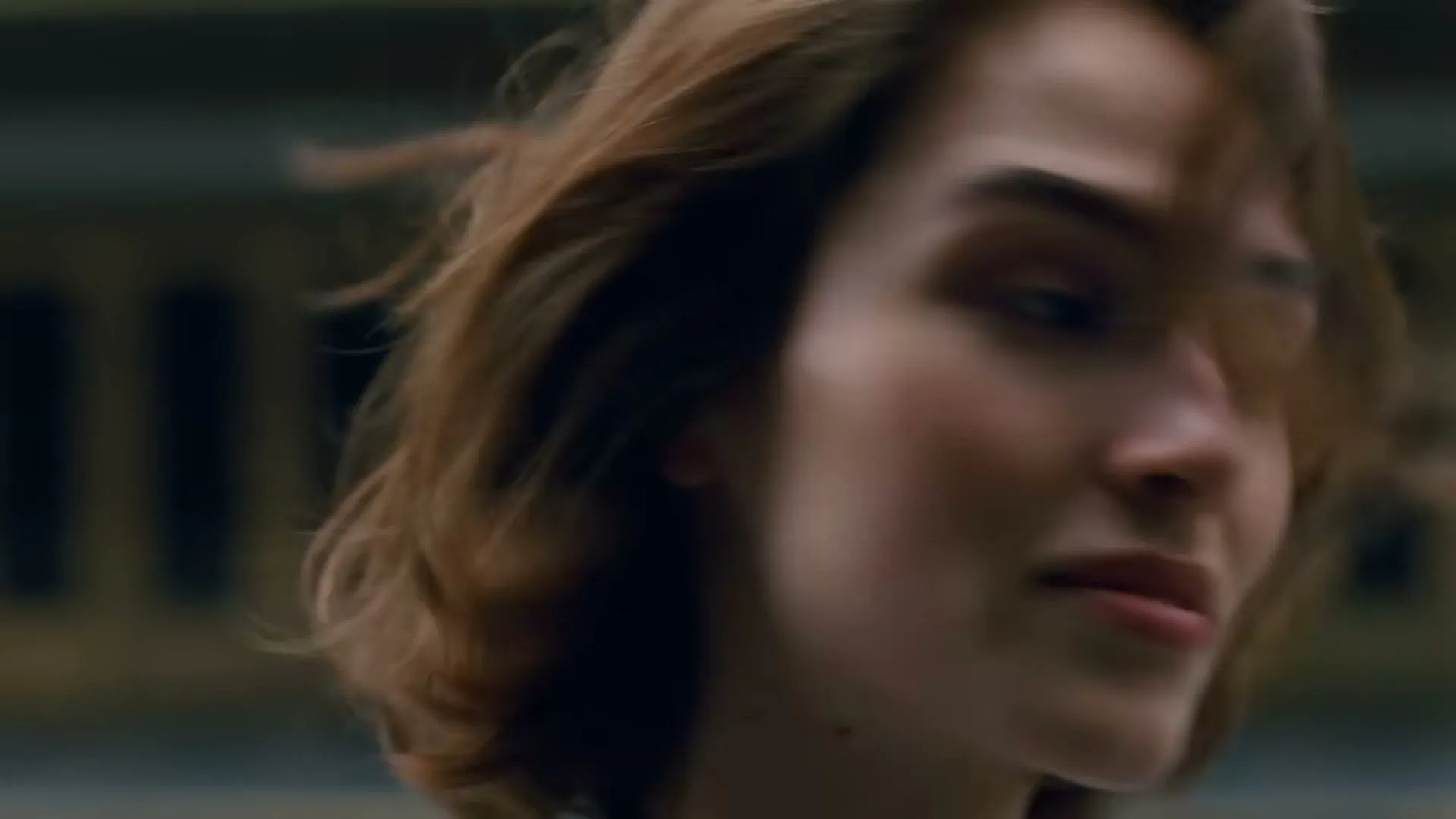 A young woman with brown hair looks to her right as her hair blows in the wind, with a blurred background of a building.
