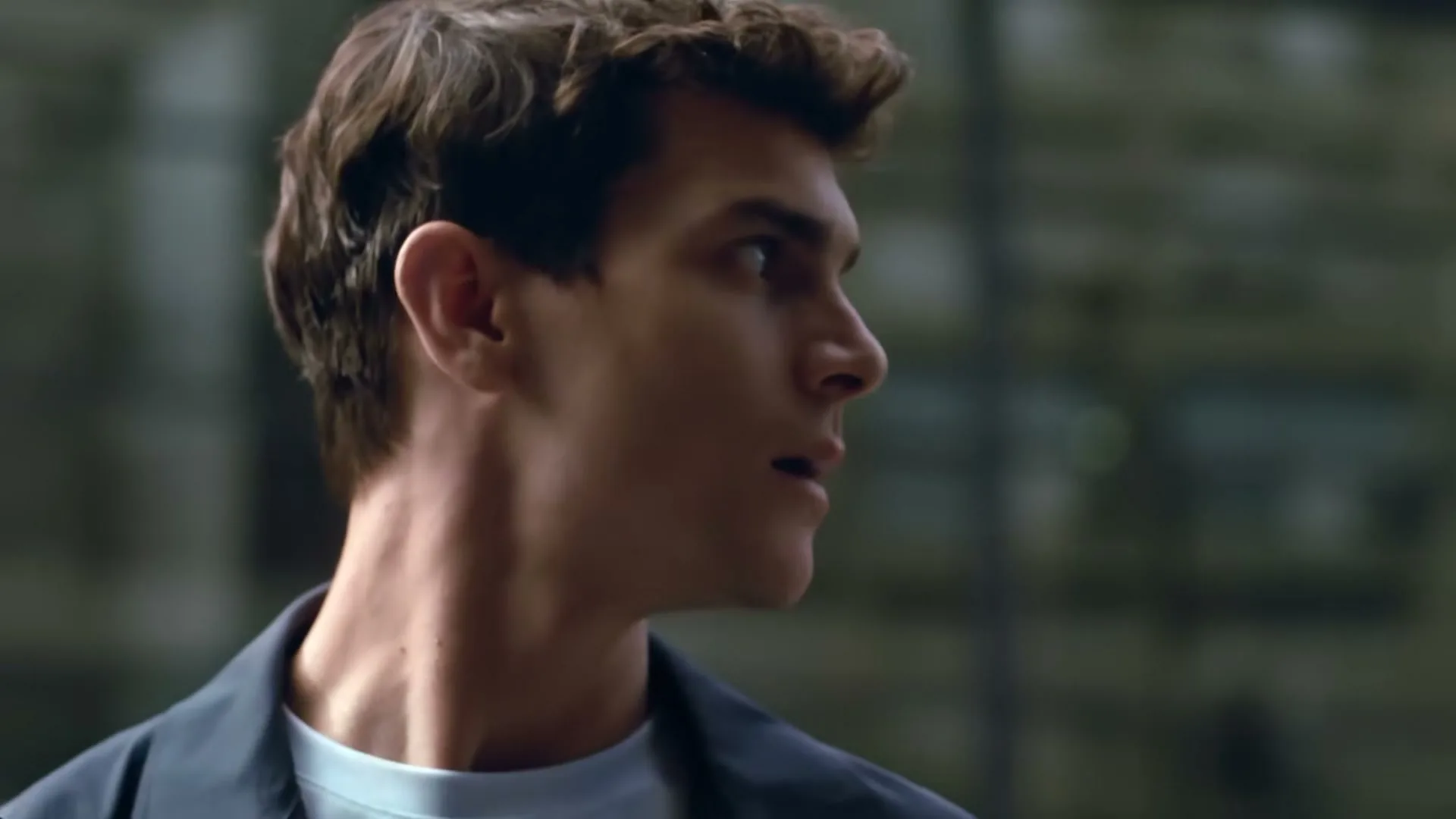 A young man with brown hair and a blue jacket looks over his shoulder in an urban exterior setting.