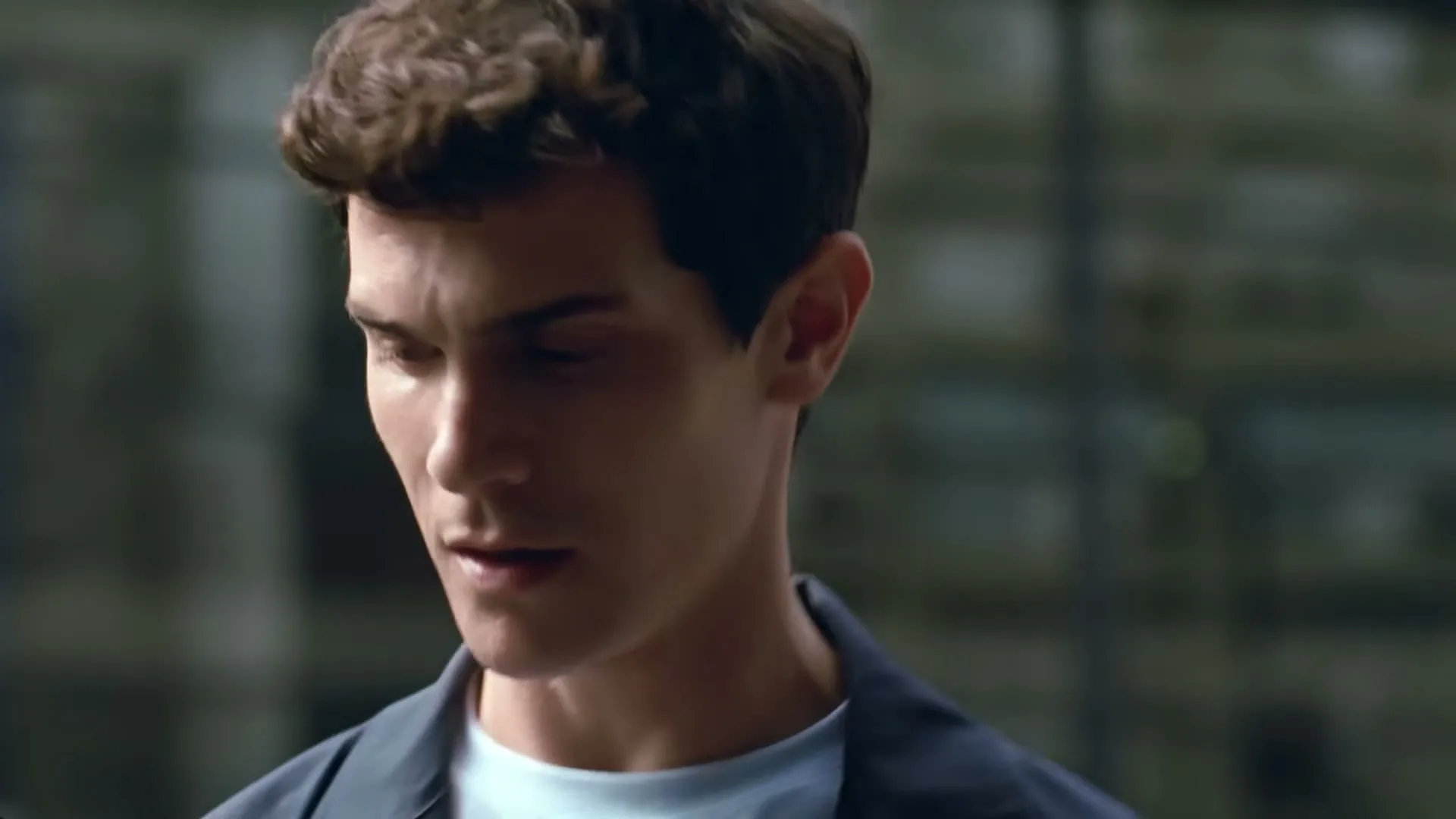 A young man with curly brown hair and a blue jacket looks down, walking in a city.