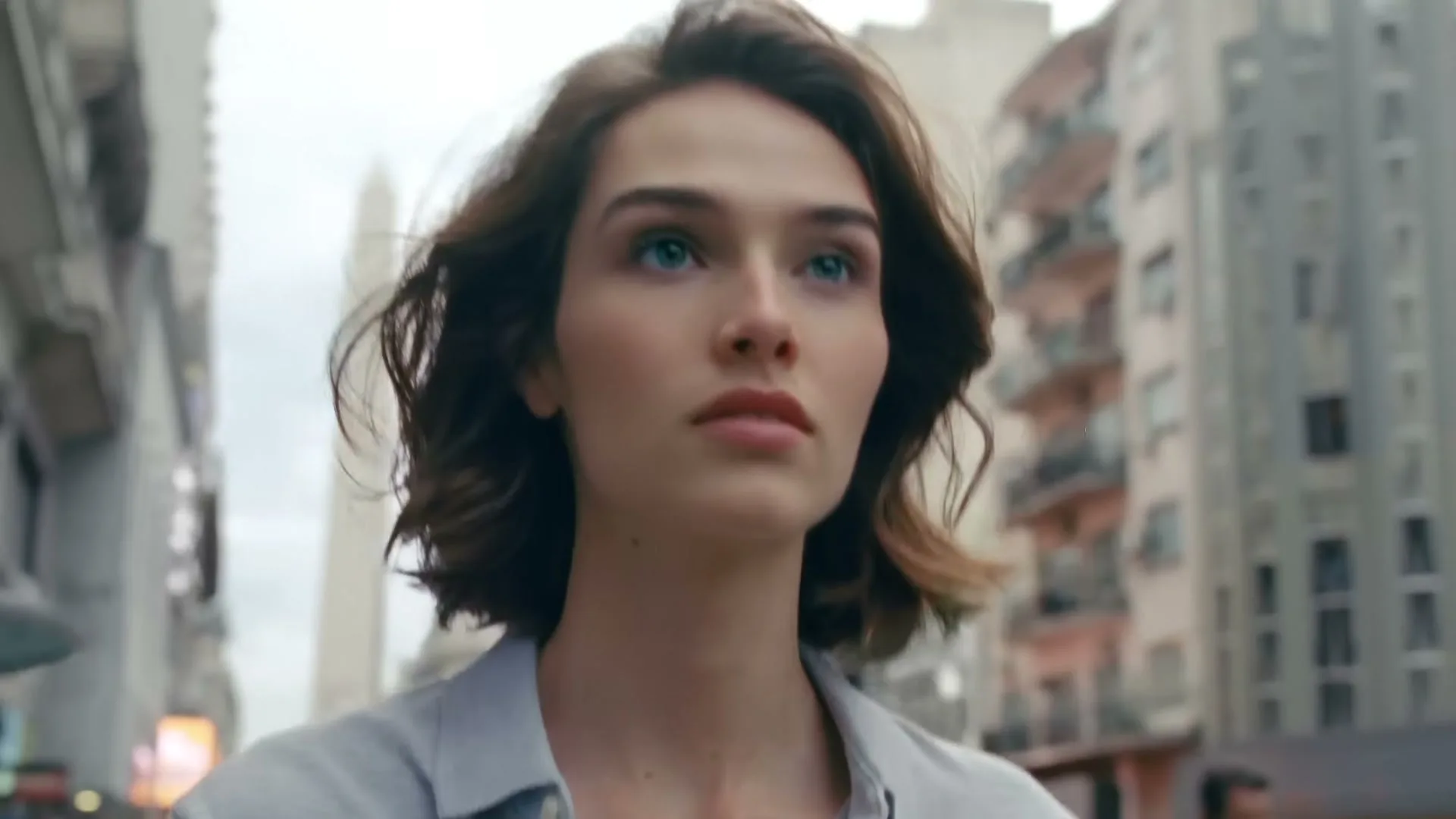A young woman with short brown hair and a light blue shirt walks through a city street, looking up with a determined expression.