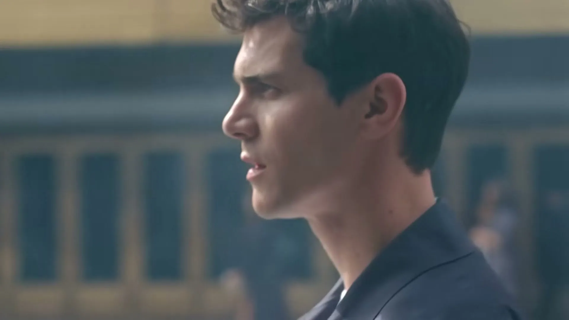 A young man with dark, curly hair and a navy shirt looks intently to the right, standing in a bustling train station.