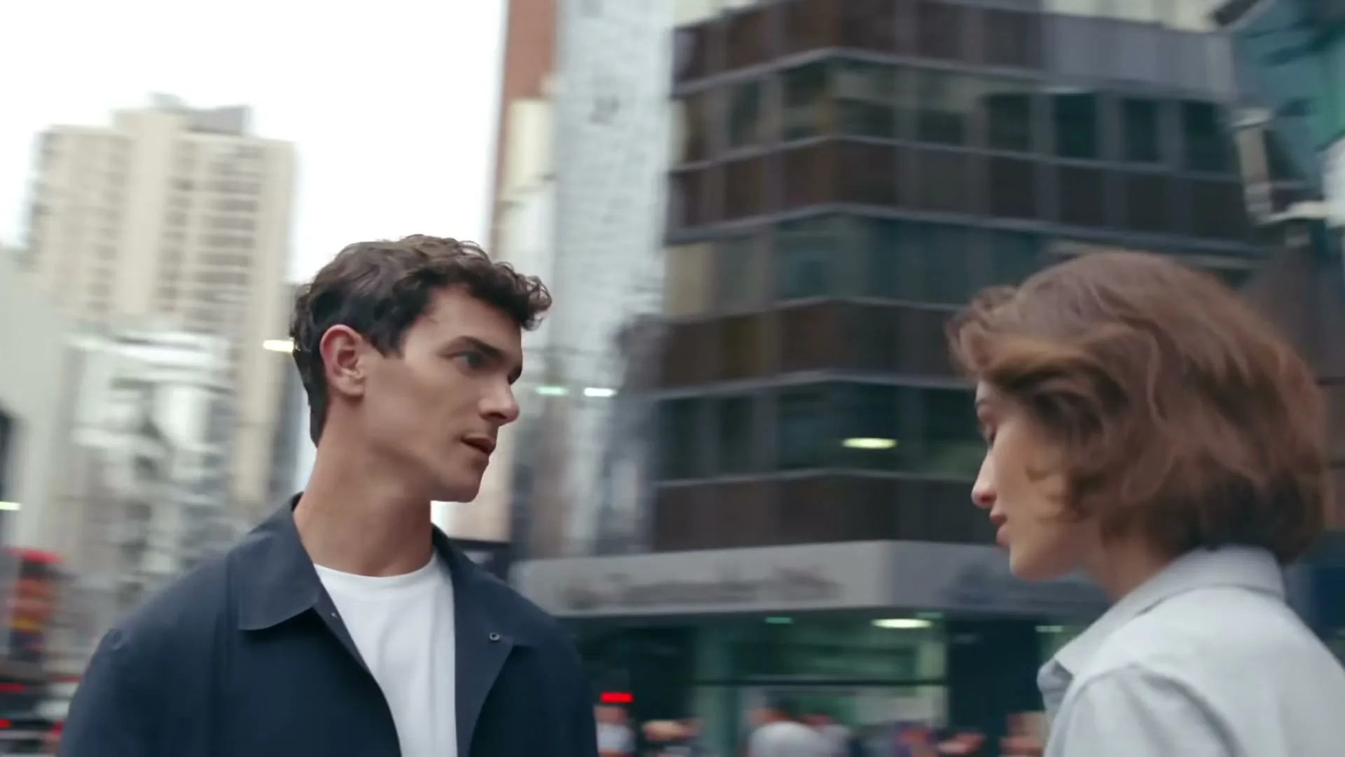 A man and a woman, both with brown hair, look at each other on a busy city street during the day.