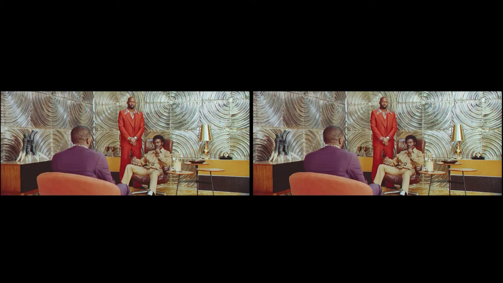 Three men in a retro-styled room, two standing and one seated, are seen from behind a fourth man in an orange chair.