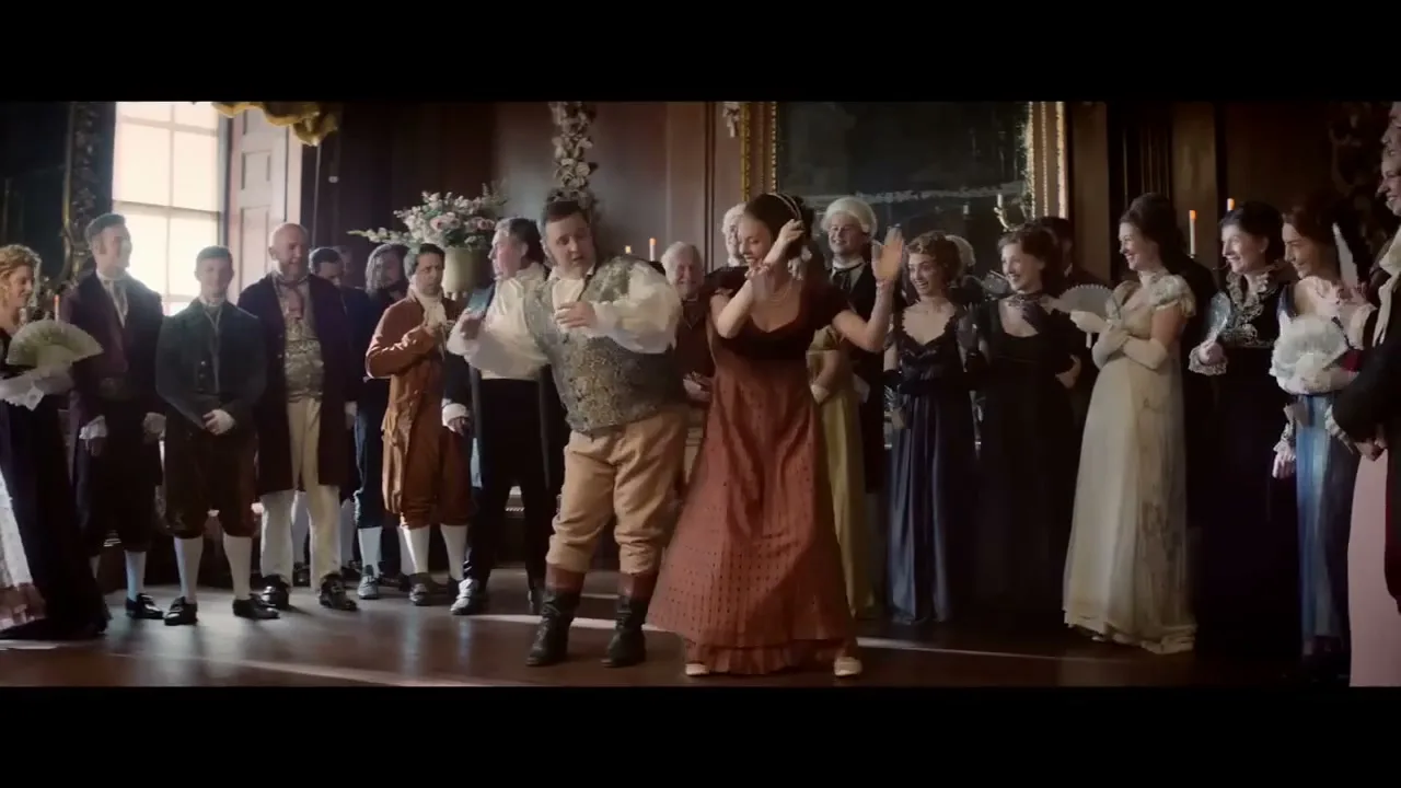 A man and woman in period costumes dance energetically in the middle of a ballroom, surrounded by other formally dressed people watching them.