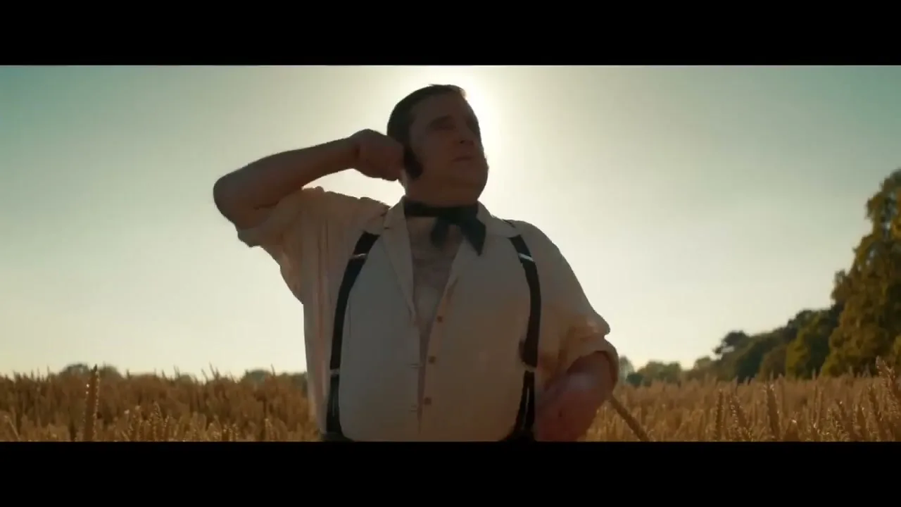 A man in period clothing stands in a wheat field, adjusting his shirt, with the sun behind him.