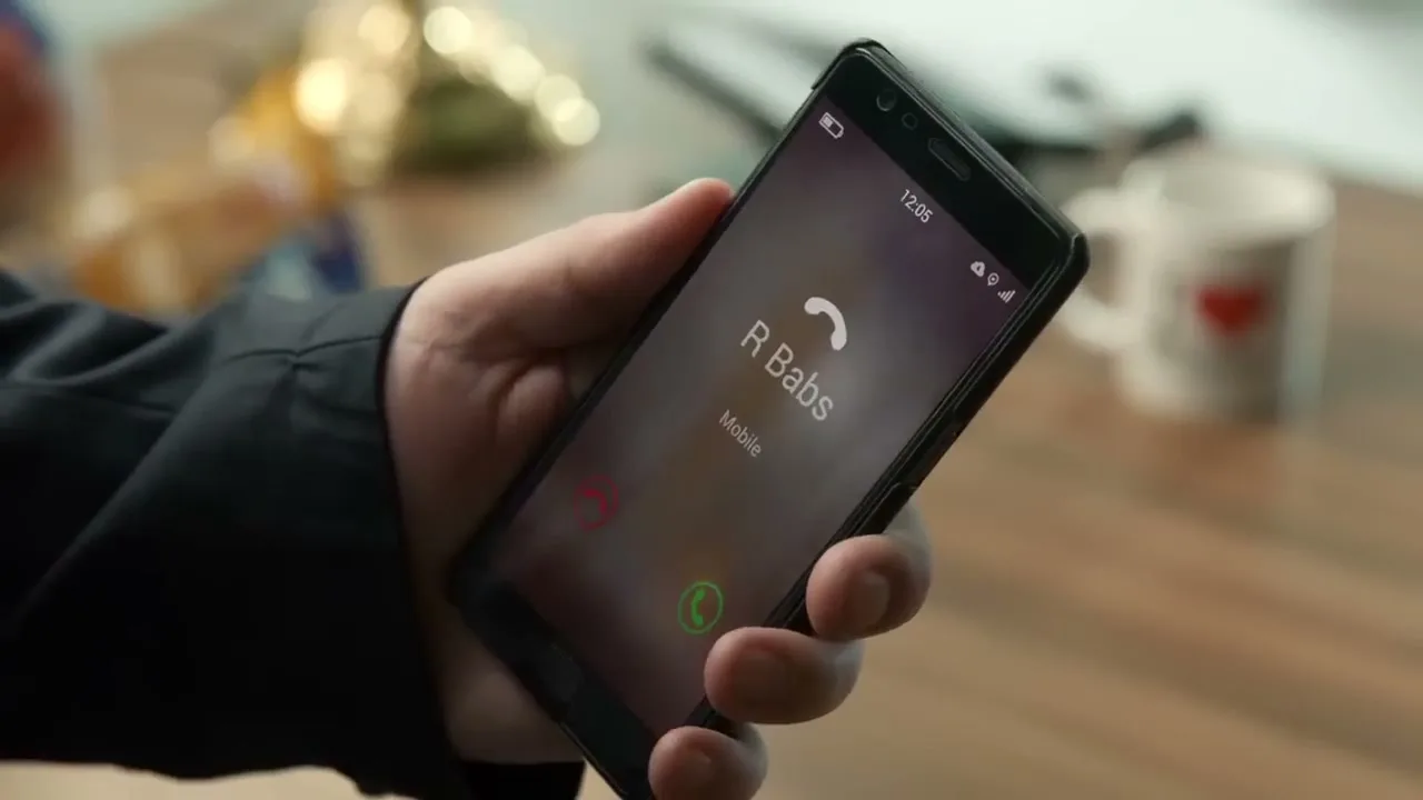 A person's hand holds a smartphone displaying an incoming call from "R Babs" on a wooden table.