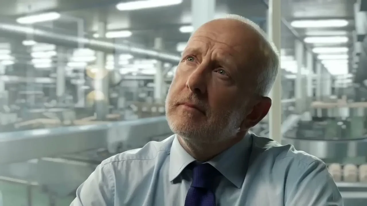 A balding man with a beard, wearing a light blue shirt and dark blue tie, looks up thoughtfully in a factory setting.