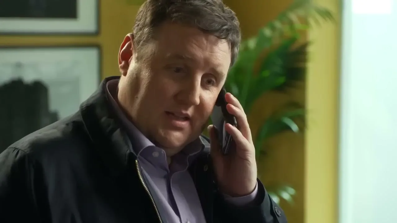 A man in his 40s, wearing a dark jacket and purple shirt, talks on a black smartphone in an office.