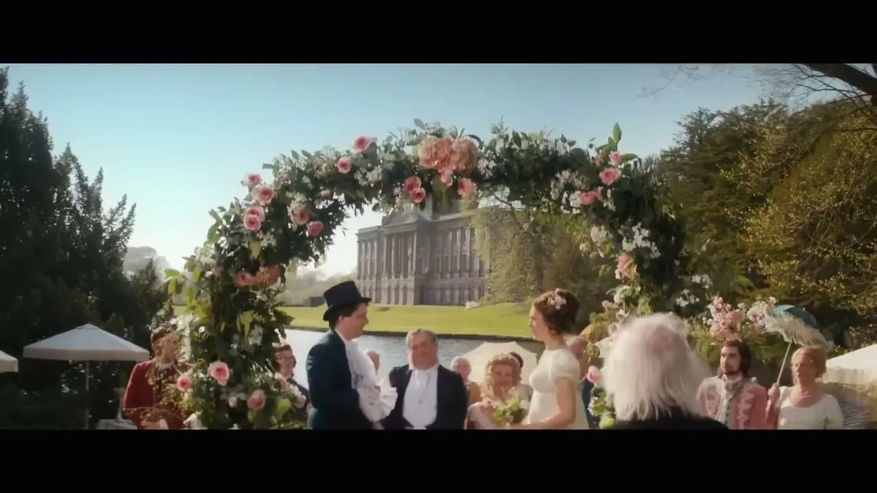 A man and woman in period clothing stand under a floral archway, getting married outdoors at a grand estate by a lake.